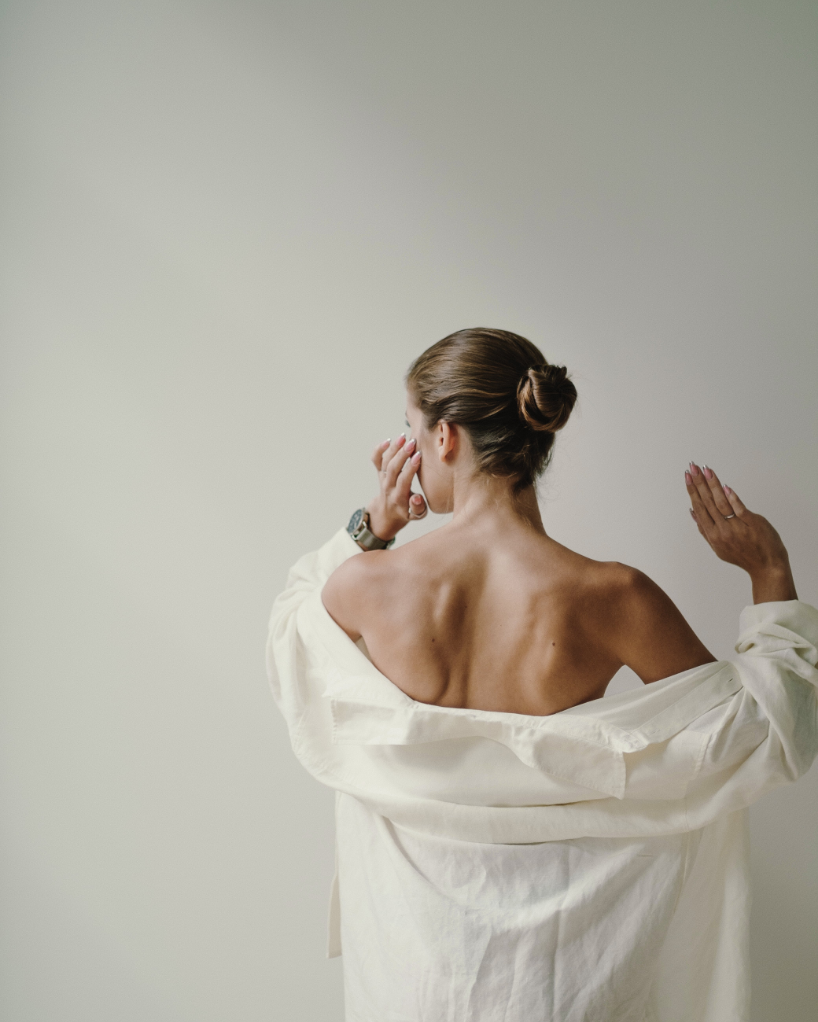 A woman with brown hair styled in a bun, standing with her back to the camera, wearing a white shirt draped off her shoulders, in front of a plain neutral background.