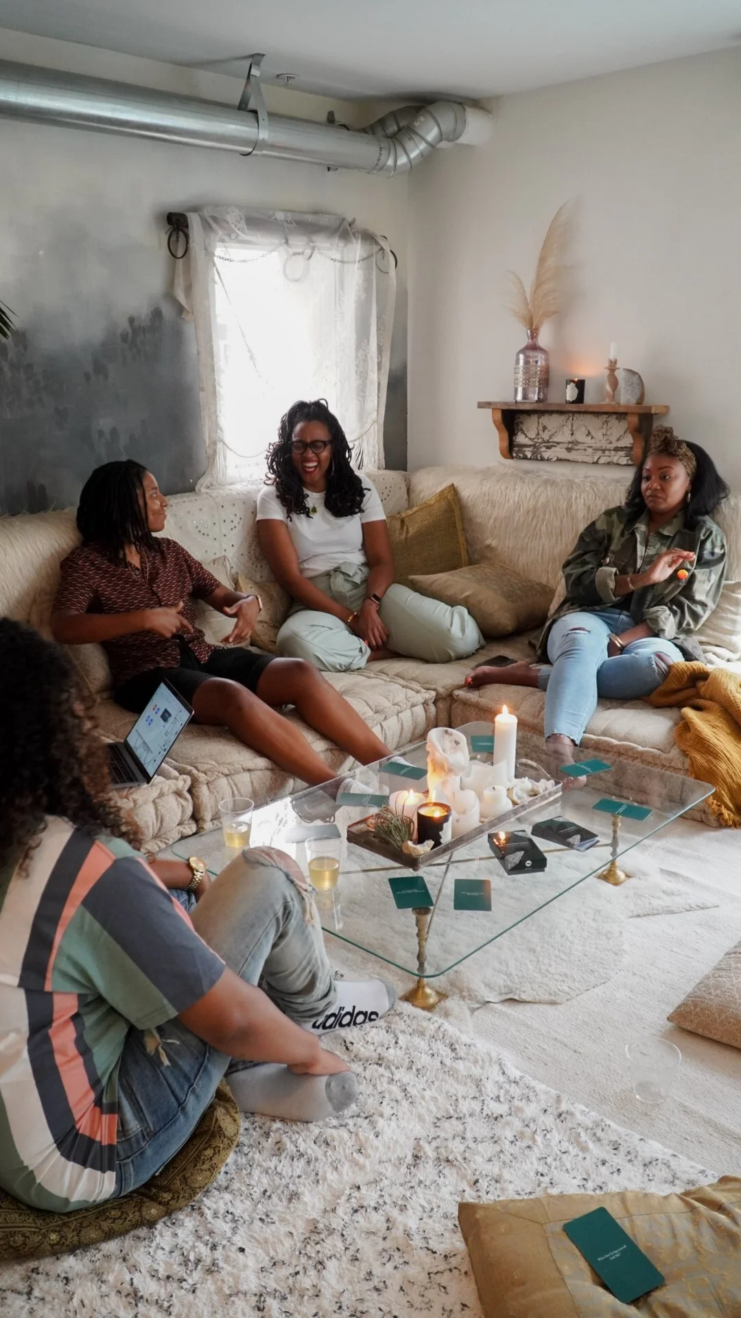 Group of five friends sitting on a beige couch in a cozy living room, enjoying conversation with candles on a glass coffee table.