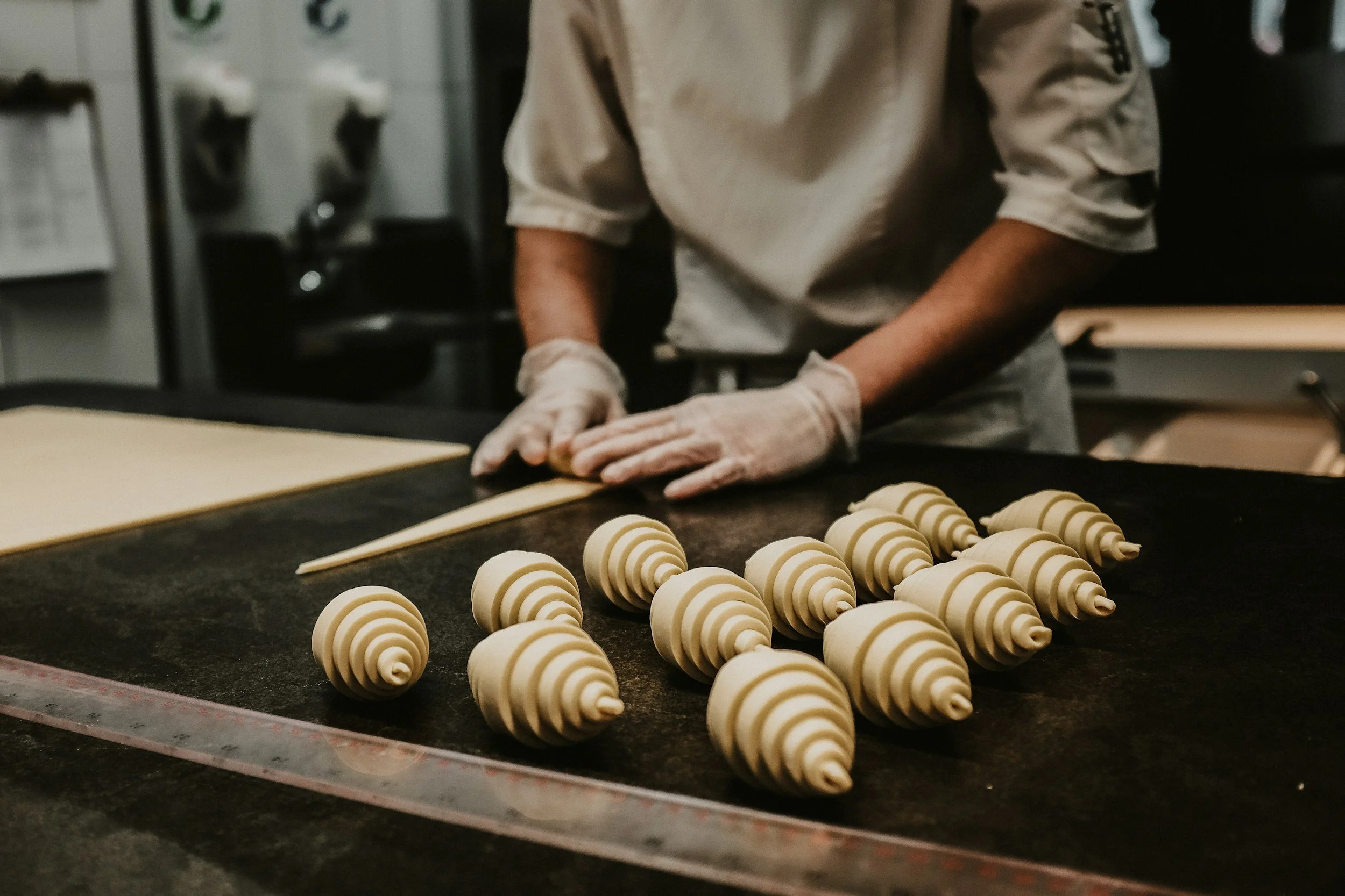 A person wearing a beige shirt and white gloves is shaping dough on a work surface with rows of spiral-shaped dough pieces.
