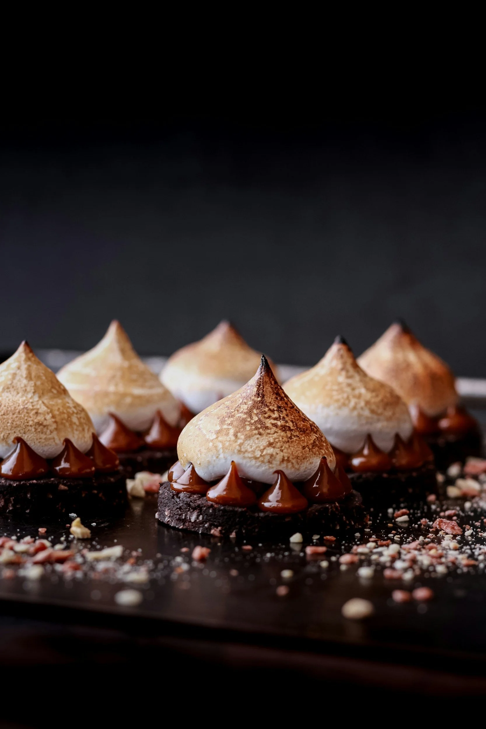 Close-up of chocolate desserts with toasted meringue peaks, chocolate drip, and crushed nuts on a black surface.