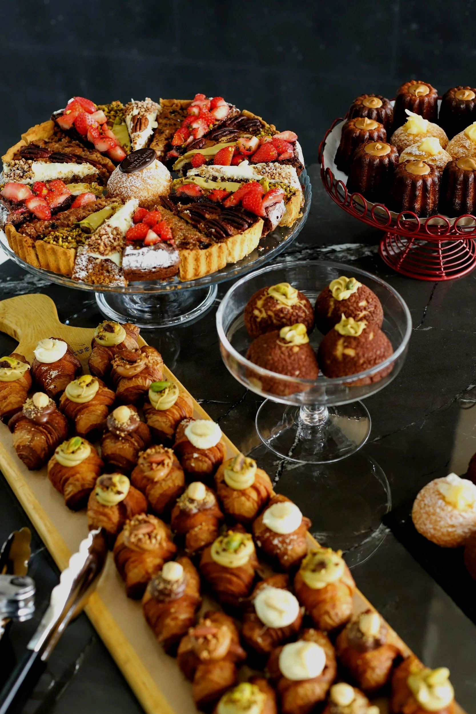 Various desserts including a large fruit tart, chocolate and vanilla bundt cakes, small pastry balls with yellow topping, arranged on dessert plates and stands on a dark marble table.