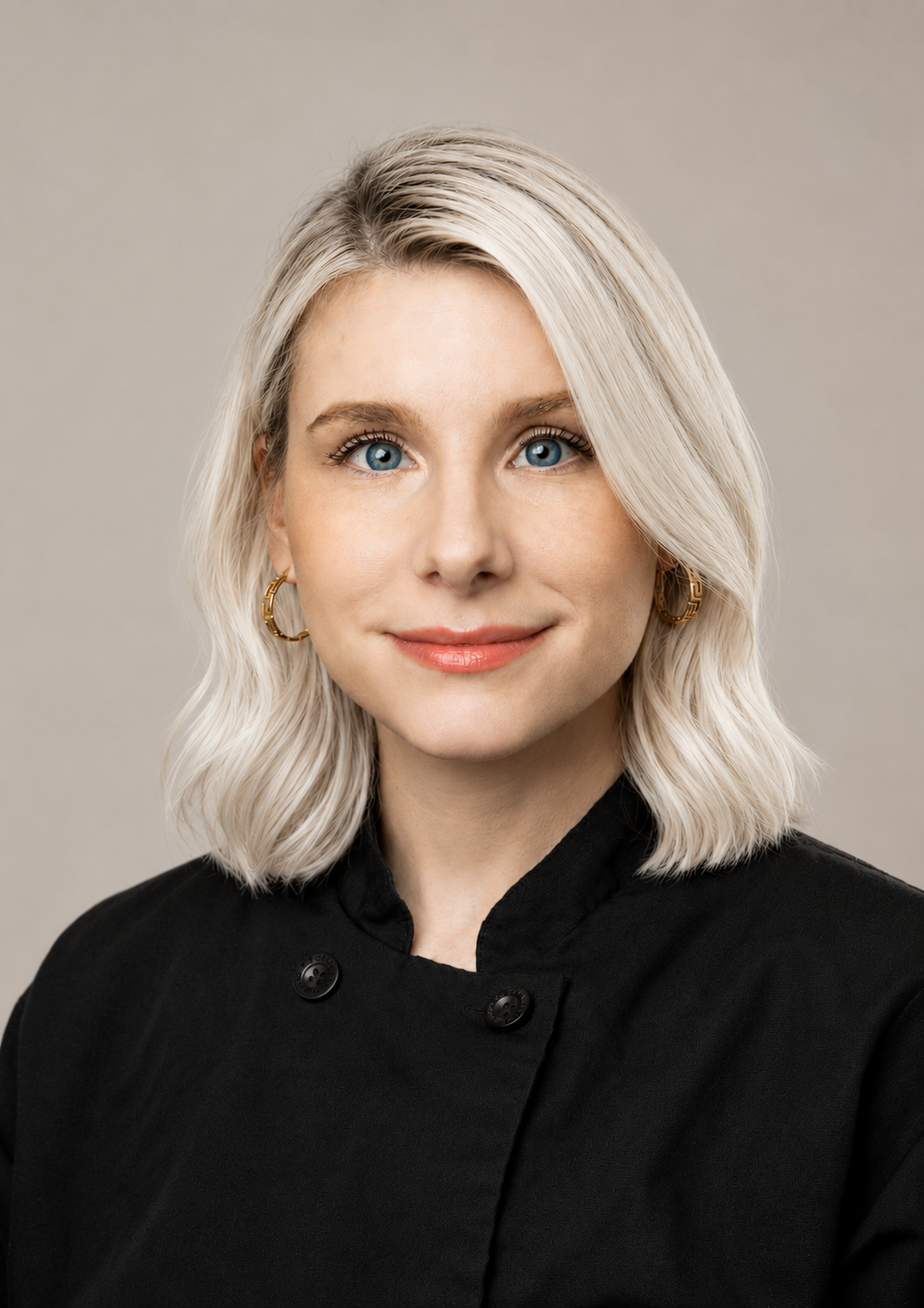 Portrait of a woman with blonde wavy hair, blue eyes, wearing gold hoop earrings and a black chef's jacket, smiling softly.