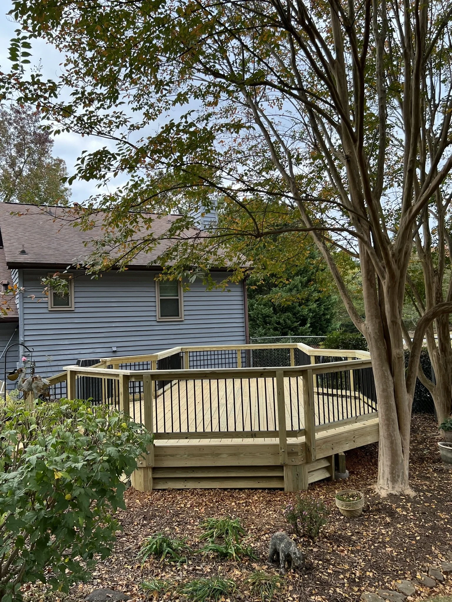 A new wooden deck with black metal railings is built around a large tree in a backyard, with a blue house in the background and trees around.
