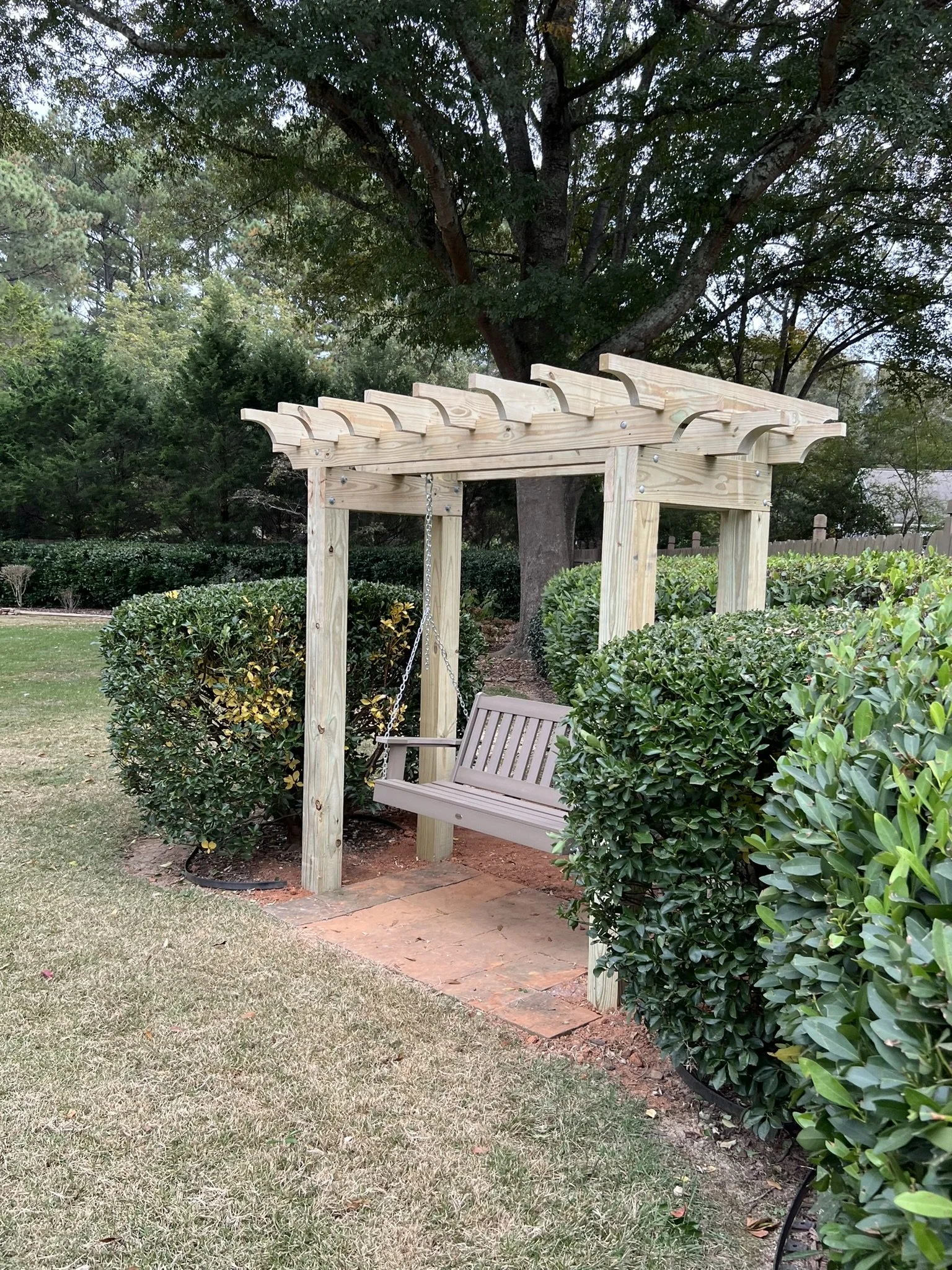 Wooden backyard swing under a pergola, surrounded by green bushes and trees