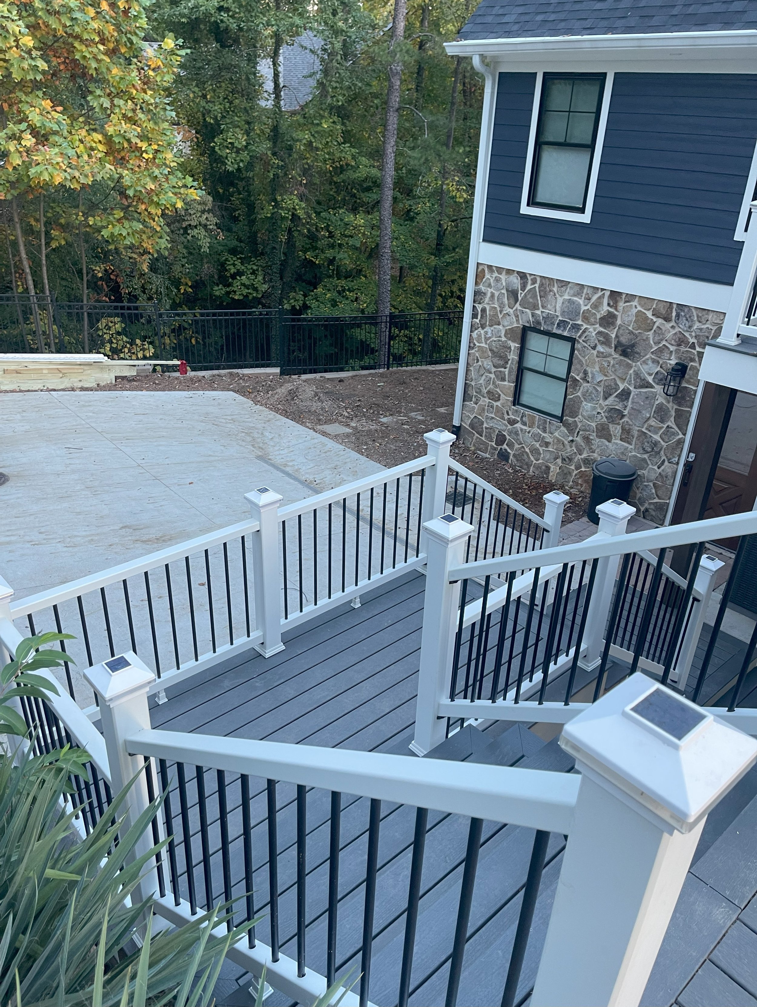 View of a backyard with a modern wooden deck with black and white railings, adjacent to a house with blue siding and stone accents. There is a partially finished concrete patio and a wooded area in the background.