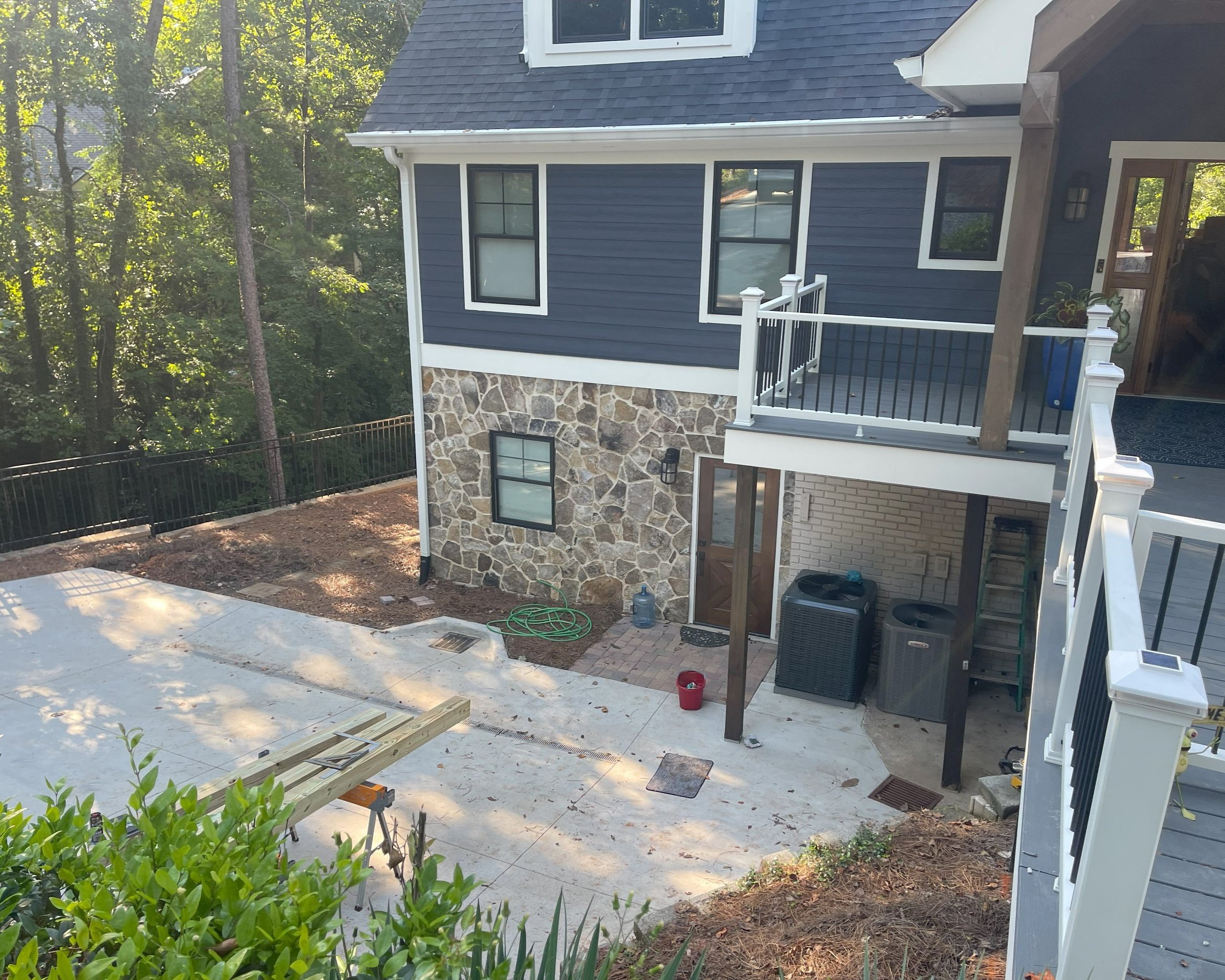 Backside of a two-story house under construction with a patio area, surrounded by trees, featuring dark blue siding, stone accents, white trim, and a balcony with black railing.