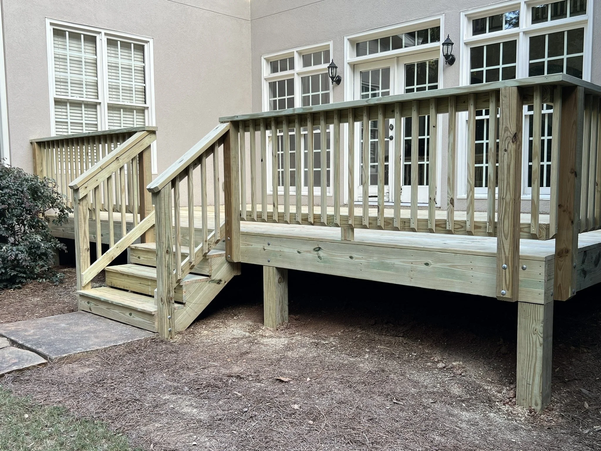 Wooden deck with stairs and railing attached to the back of a house, with large windows and a sliding door, and some shrubs nearby.