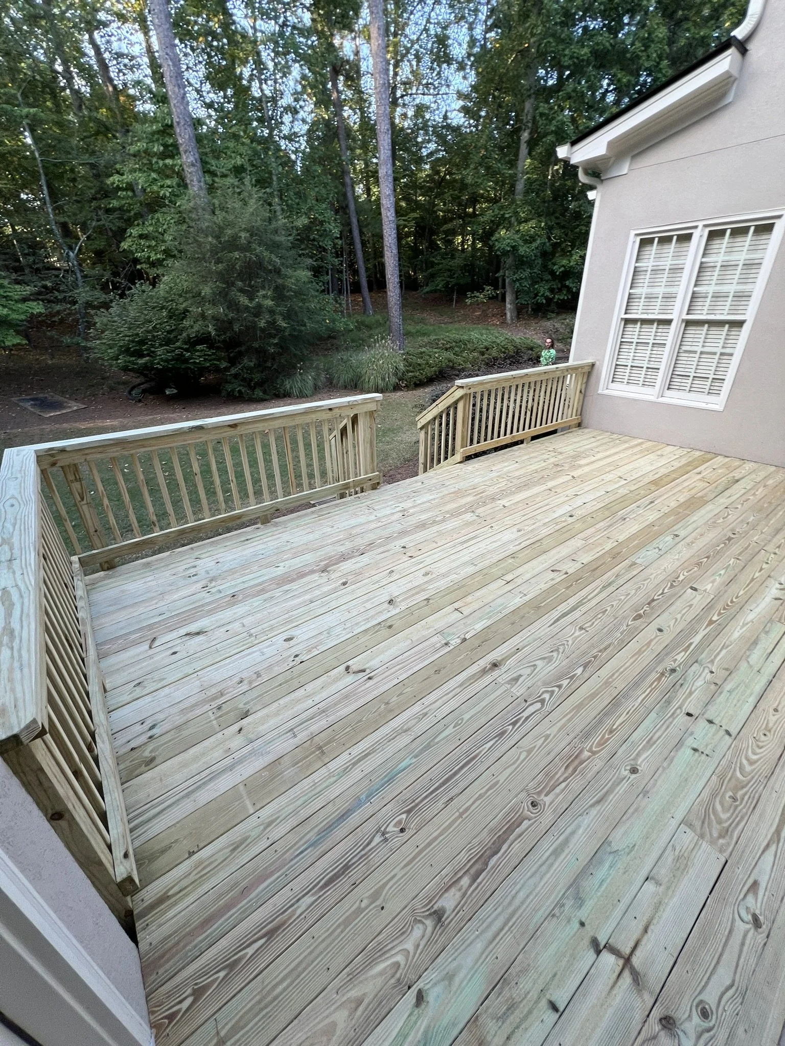 Newly built wooden deck attached to a house, overlooking a backyard with trees and greenery, with a person standing near the treeline.