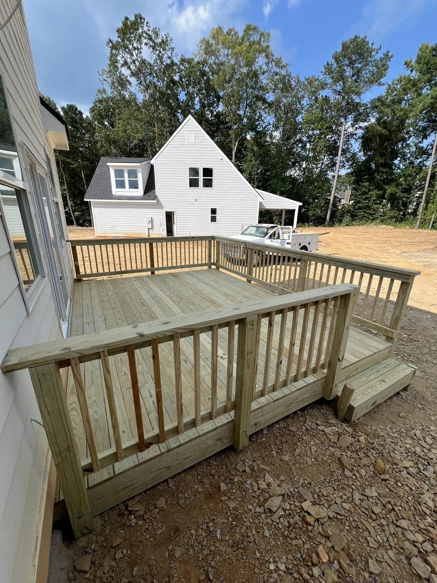 Newly built wooden deck attached to a house with steps leading down to dirt, with a background of a house, trees, and a truck.