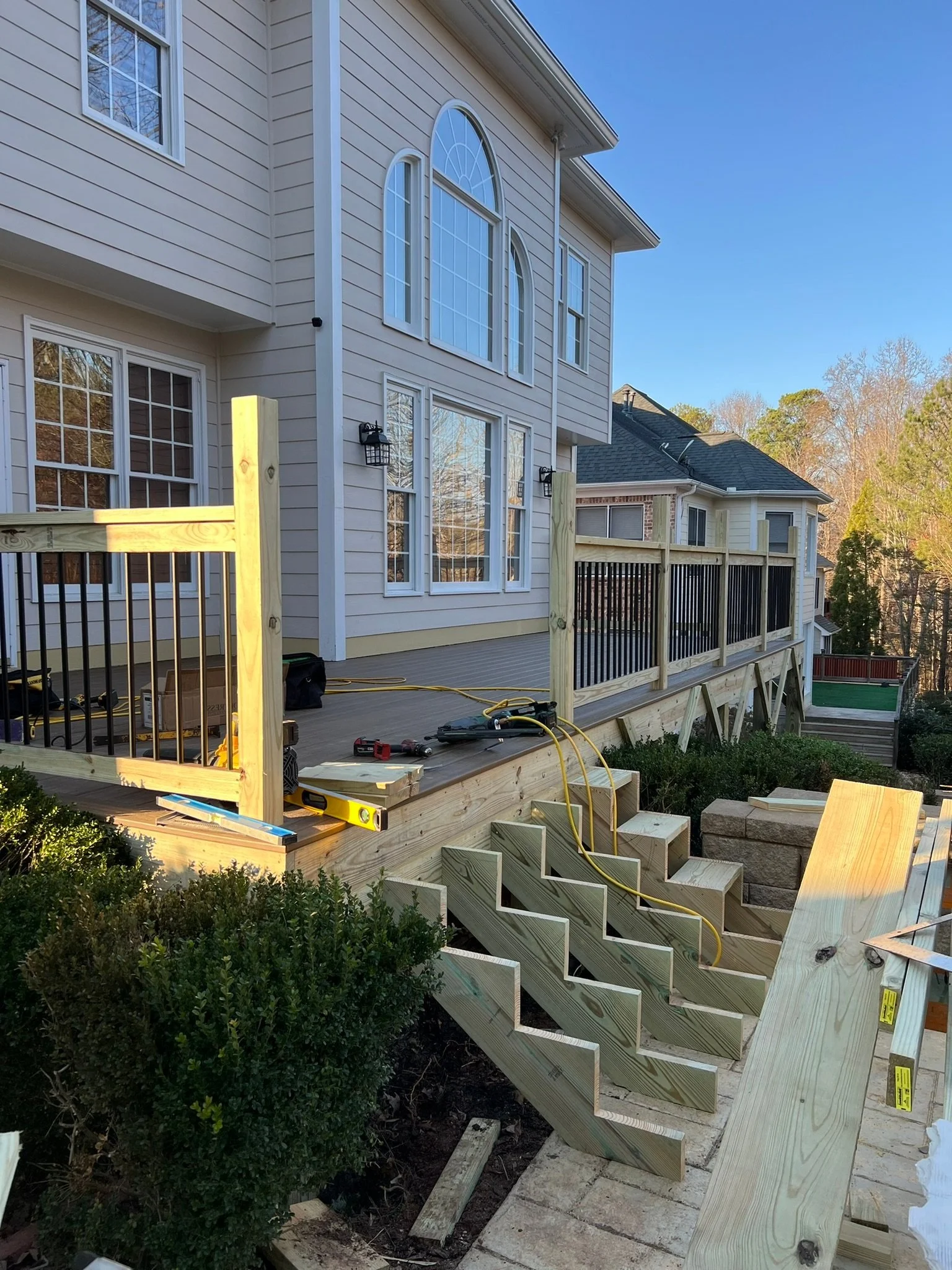 Construction of a wooden deck attached to a beige house with large windows, stairs, and black railings, on a sunny day.