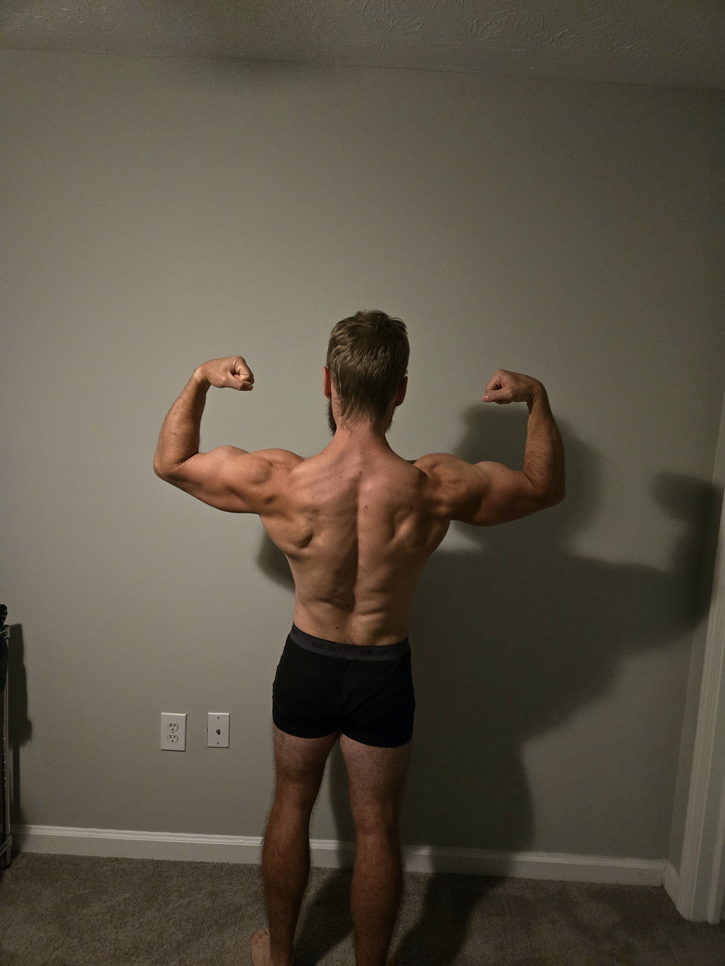 Back view of a muscular man with brown hair and beard, flexing his arms in a room with beige walls and electrical outlets, wearing black underwear.