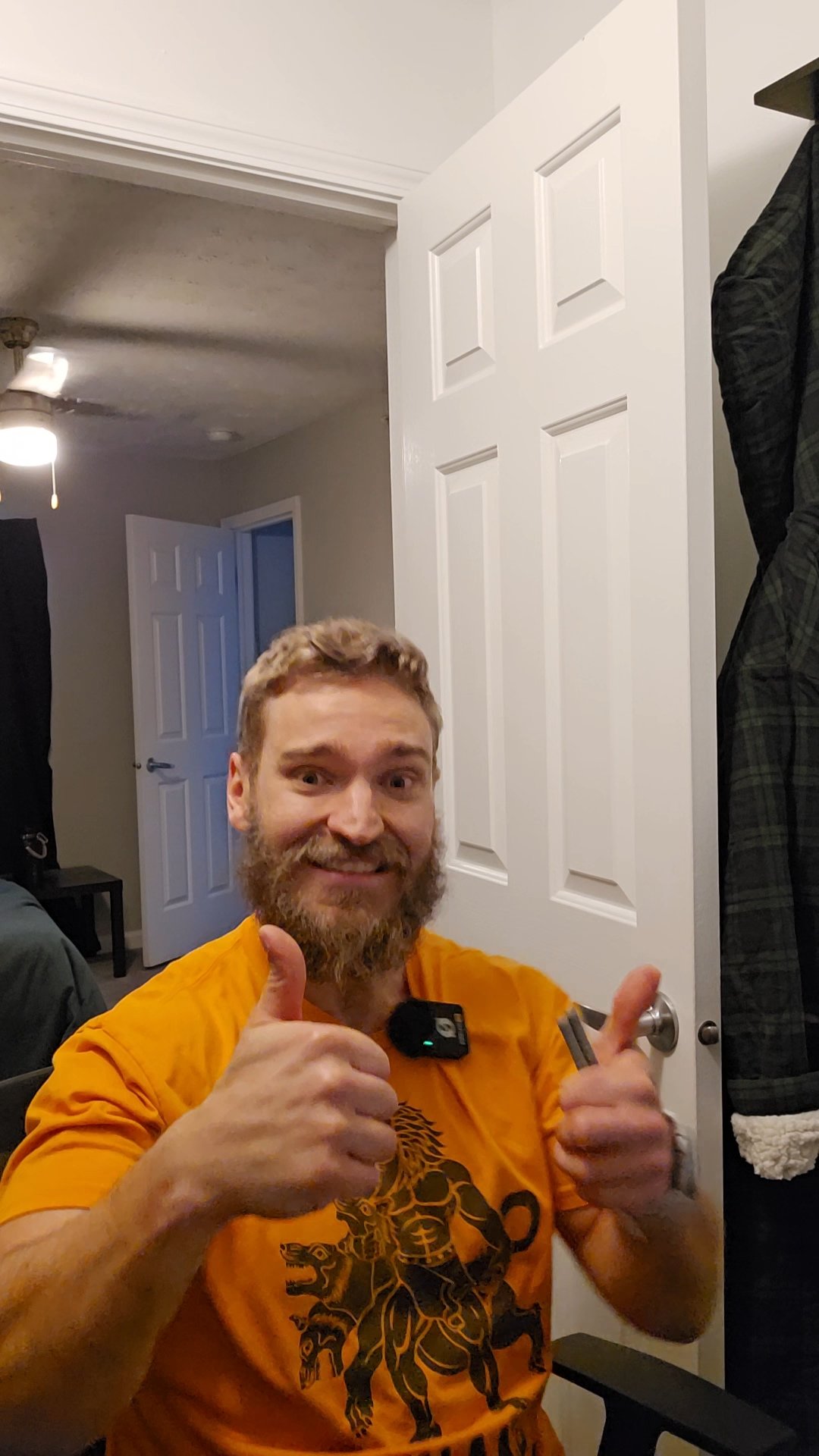 Man with a beard wears an orange shirt with a lion graphic, giving two thumbs up while smiling inside a room with a partly open door.