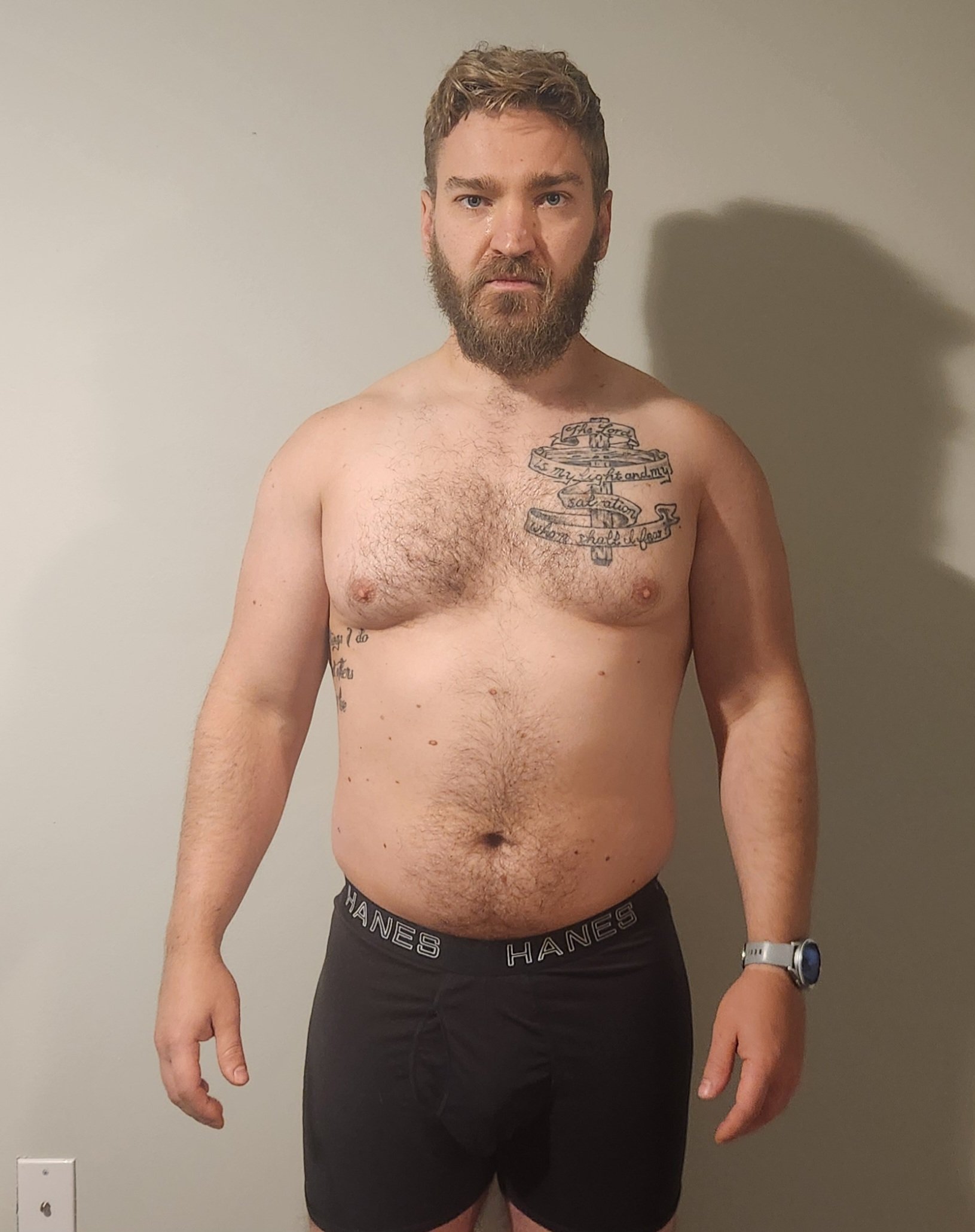 A shirtless man with a beard and short hair standing against a plain wall, wearing Hanes underwear and a watch, with tattoos on his chest and arm.