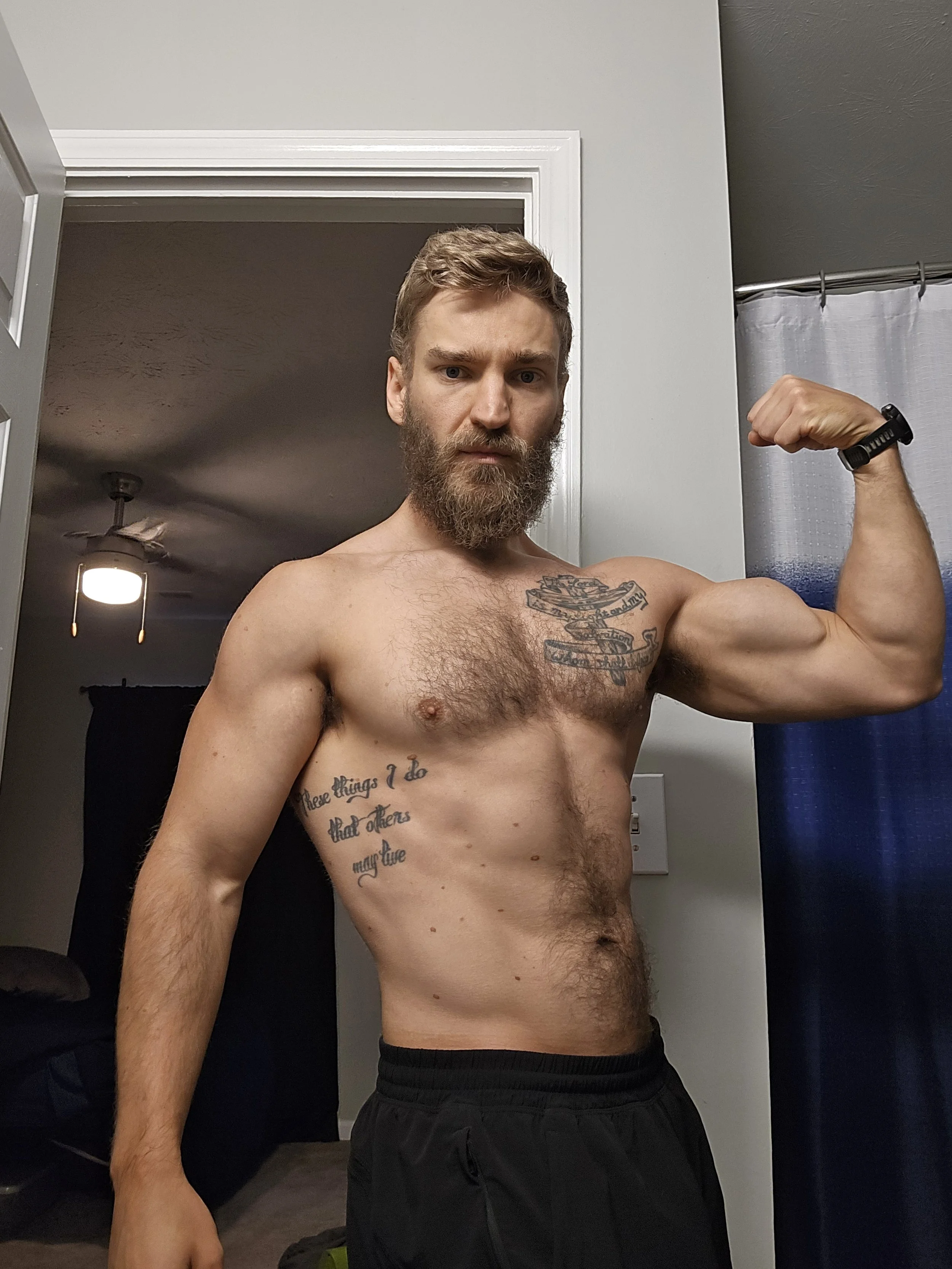 A shirtless man with a beard and tattoos flexes his right bicep in a mirror, taking a selfie in a room with a gray door, a ceiling fan, and a blue shower curtain.