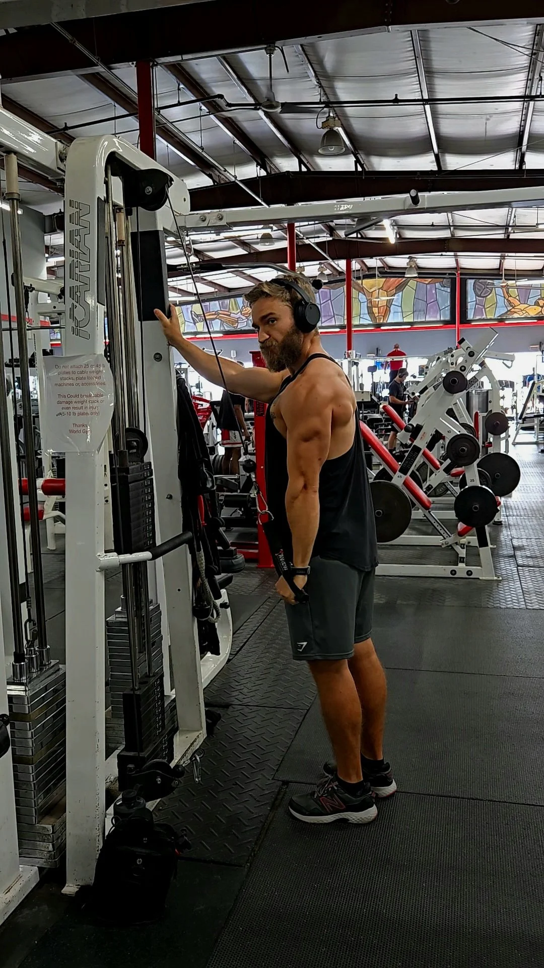 A man with a beard wearing headphones, a black tank top, and shorts, standing in a gym, leaning on a piece of exercise equipment.