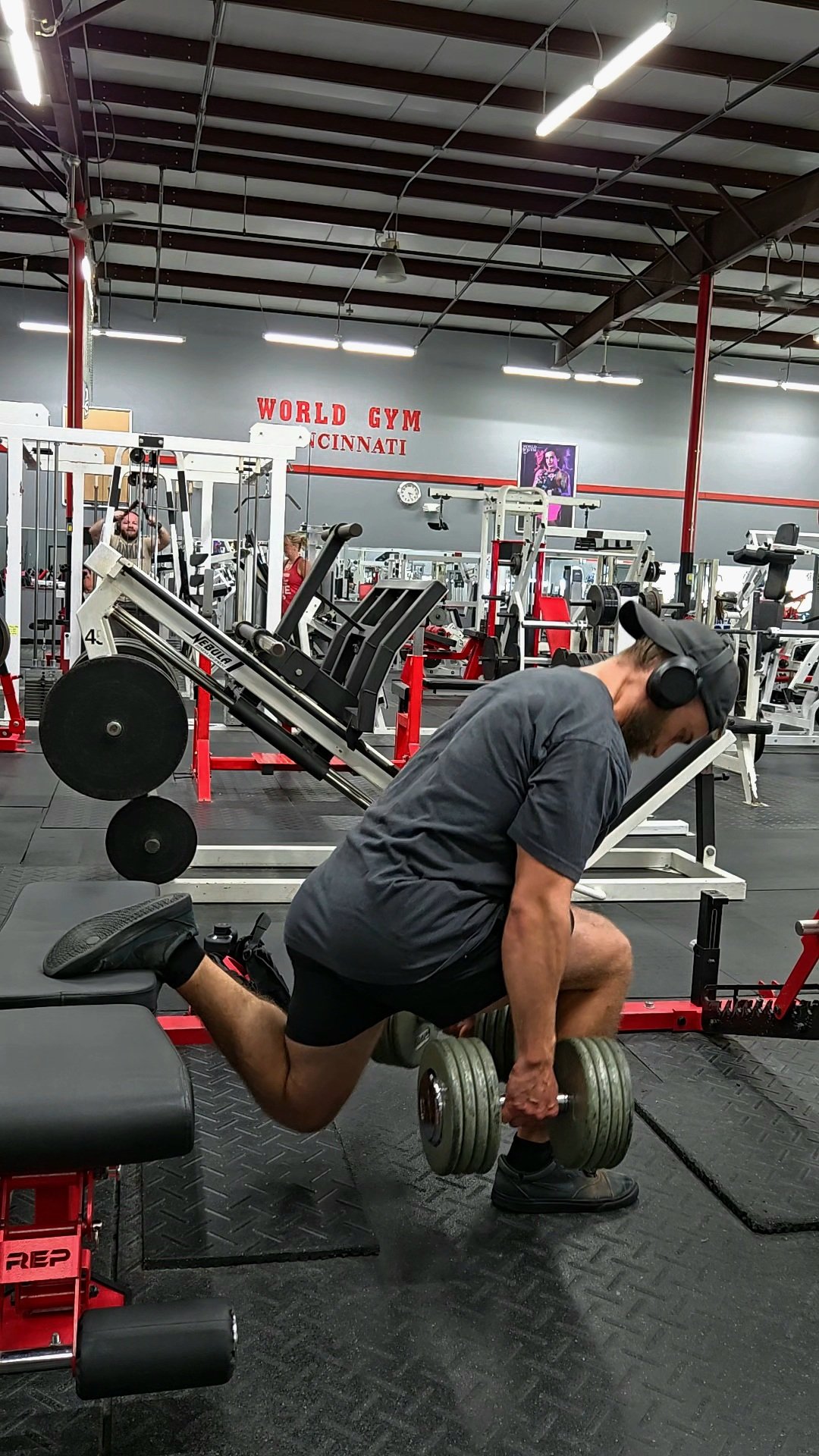 A man lifting dumbbells with one knee on a bench in a gym, wearing headphones, a gray shirt, and black shorts.