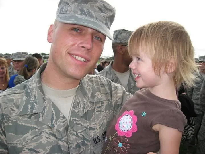 A soldier in camouflage uniform holding a young girl with blond hair, smiling and looking at each other, with other soldiers in the background.