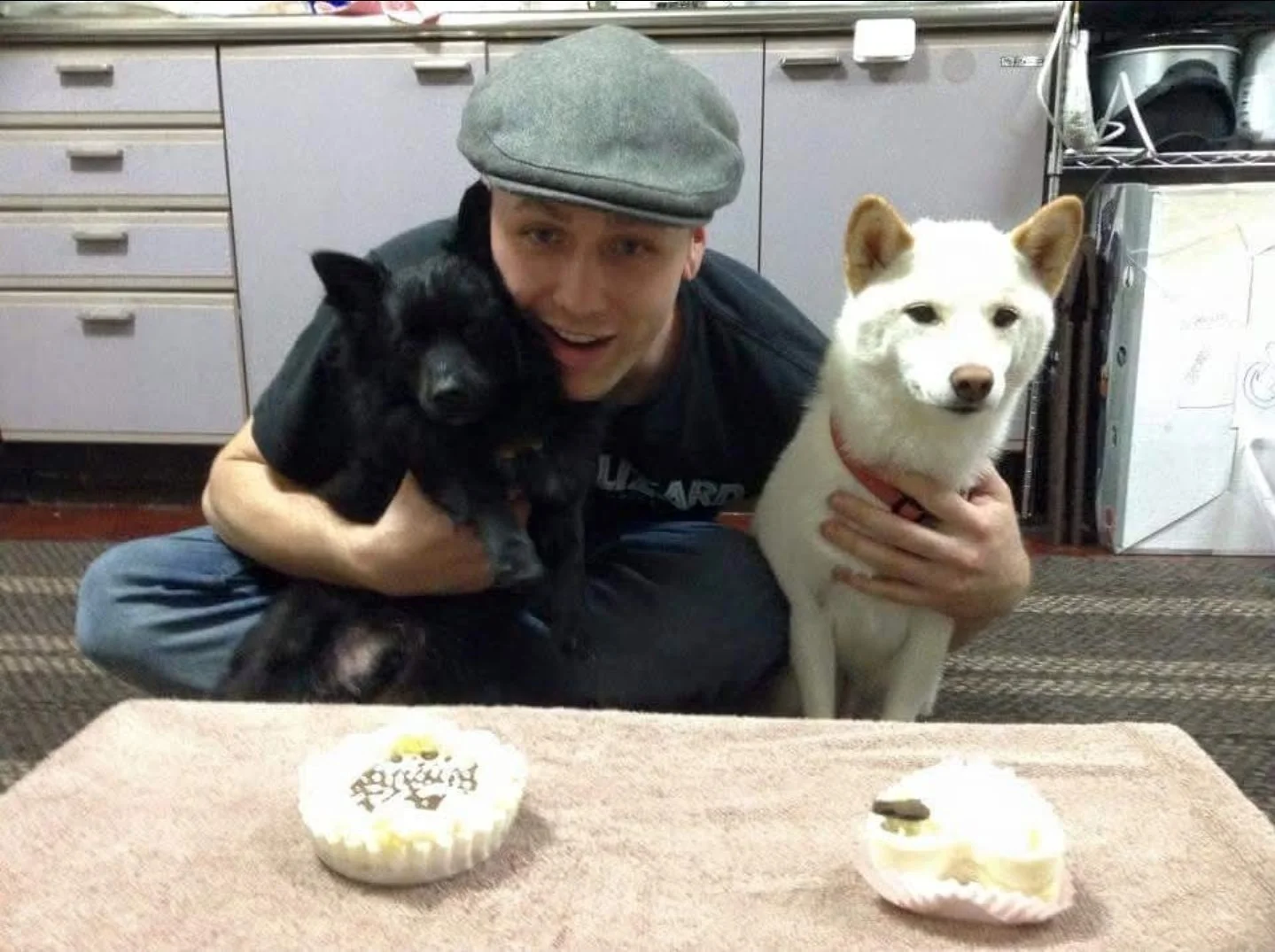 A man wearing a gray cap and dark clothing sitting on the floor, holding a black puppy in his left arm and a white dog with brown ears and a red collar in his right arm. Two cupcakes with white icing and chocolate toppings are on a pink towel in foreground, with one cupcake decorated with writing.