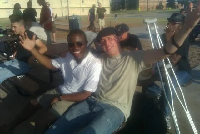 Two young men sitting on a park bench smiling and waving at the camera. One is wearing a white shirt, sunglasses, and the other a gray T-shirt. Multiple people are in the background at an outdoor event.