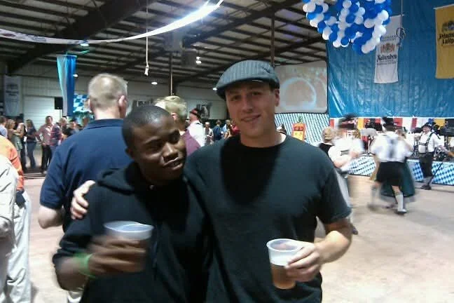 Two men standing together at an indoor event, holding drinks and smiling at the camera, with balloons and people in the background.
