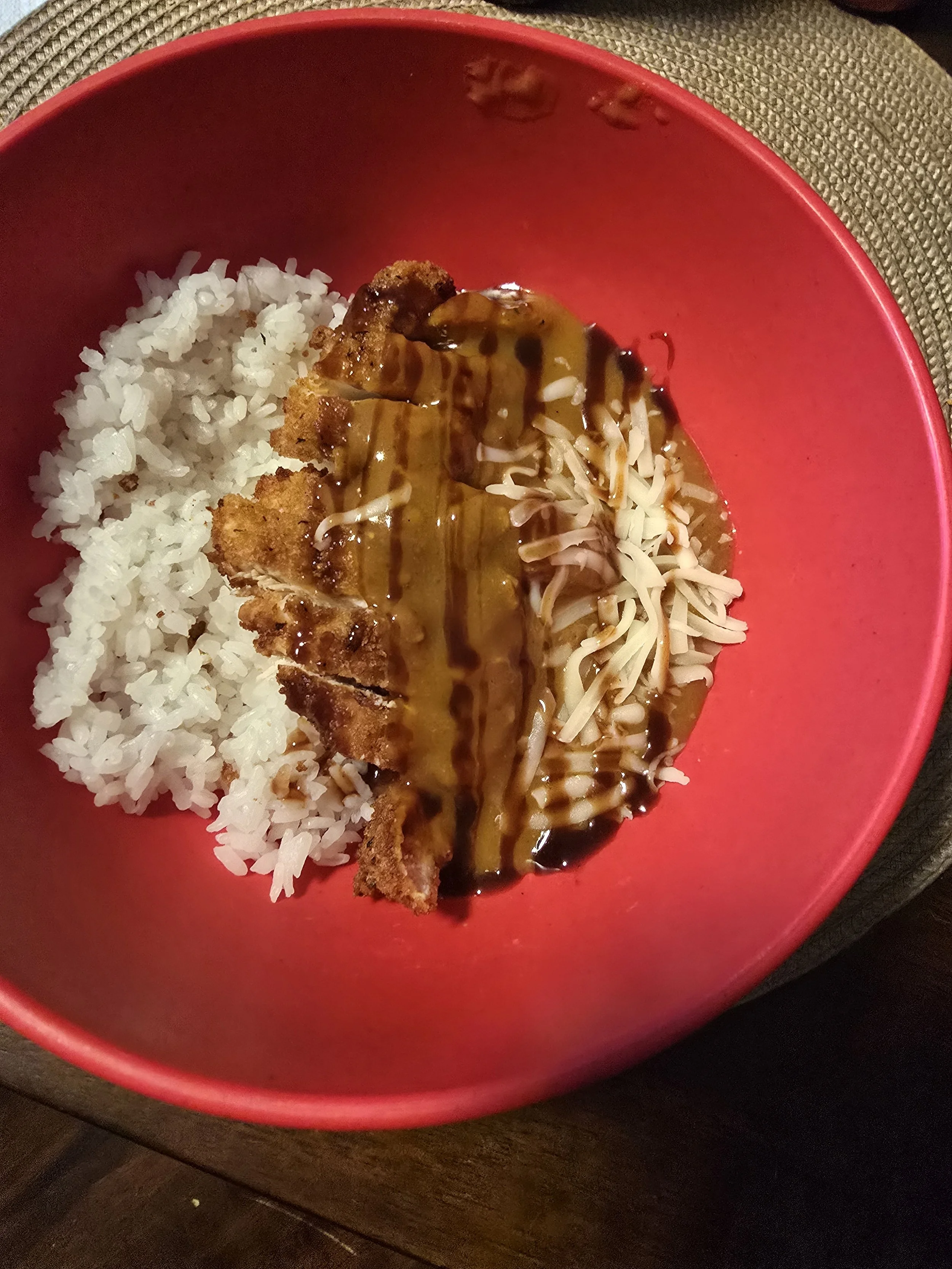 A red bowl containing white rice, breaded fried chicken with gravy, shredded cheese, and layered sauces.