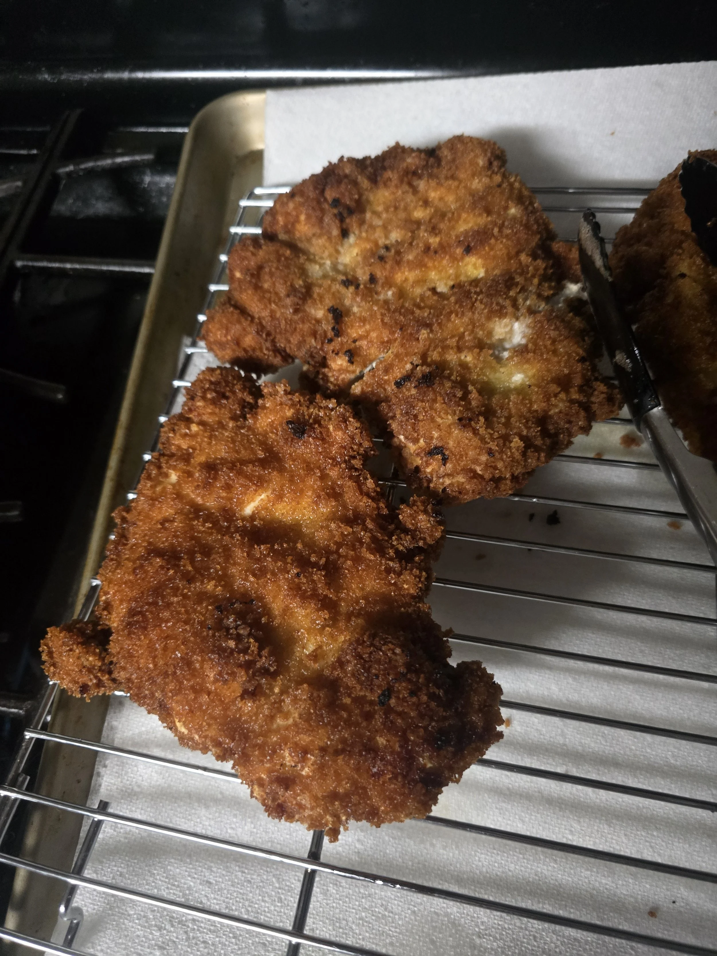 Two pieces of golden-brown fried fish resting on a wire cooling rack over a paper towel inside an oven.