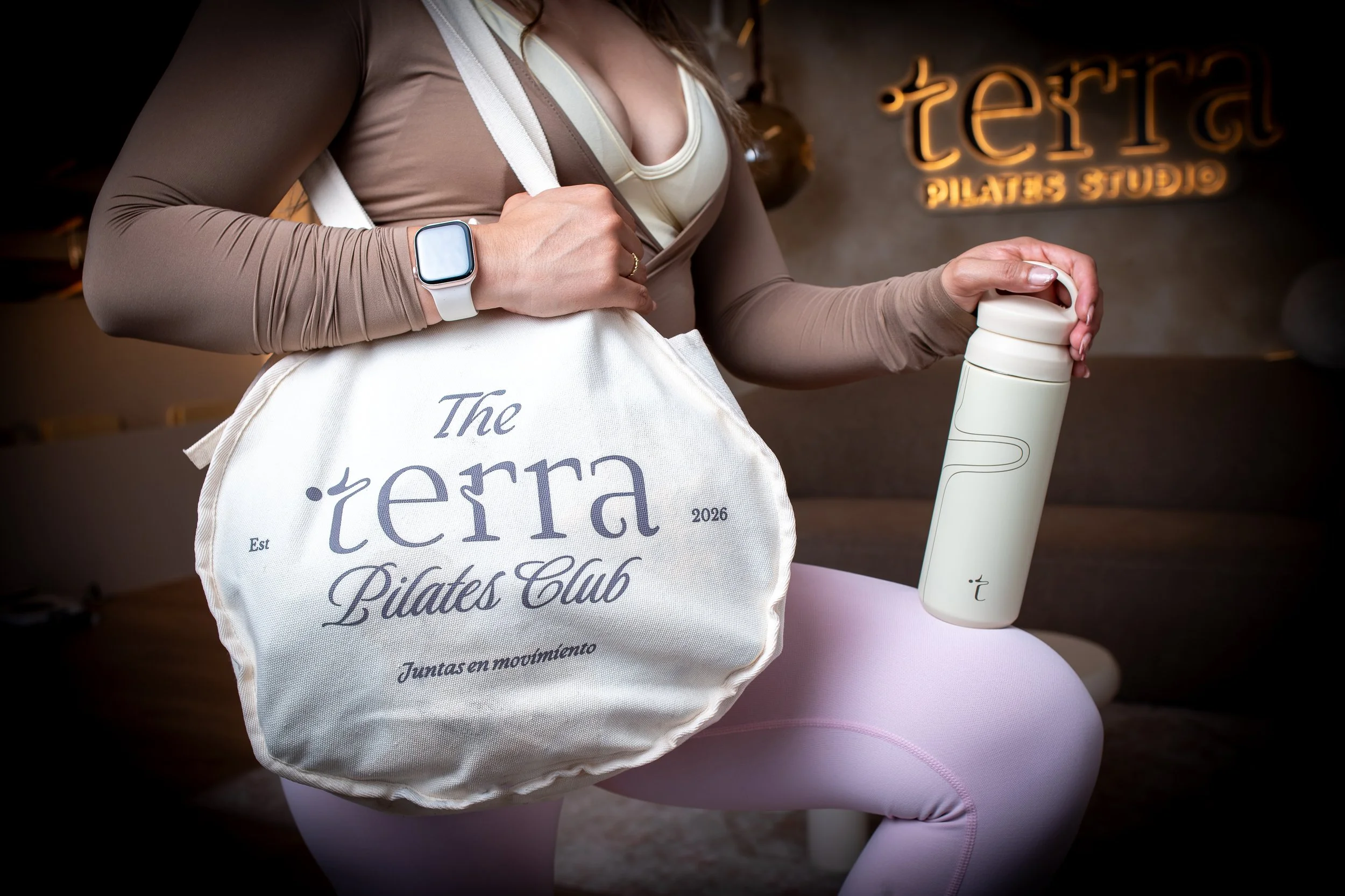 Mujer con ropa deportiva y reloj inteligente sostiene una botella blanca de agua, lleva una bolsa de tela con el logo del club de pilates y está en un estudio de pilates.