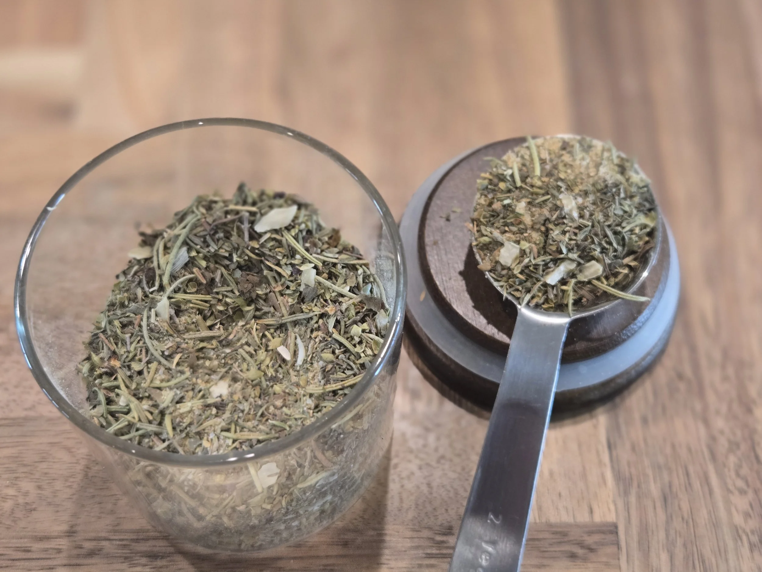Two glass containers filled with dried herbs placed on a wooden surface, with metal measuring spoons beside them.