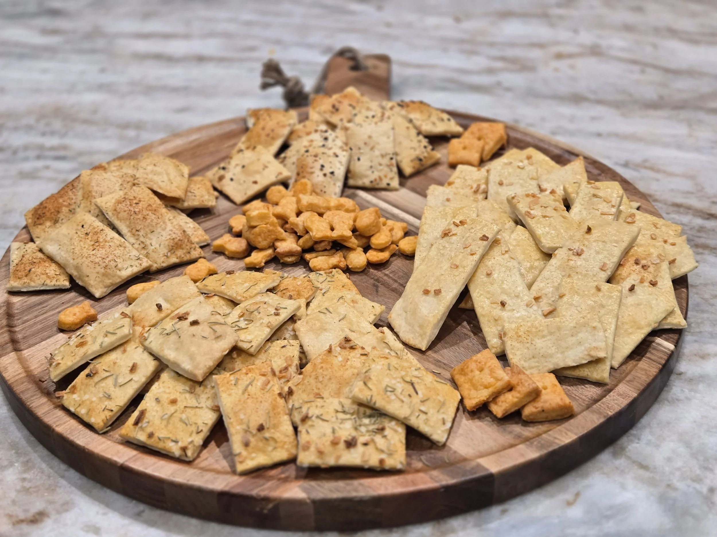 A round wooden platter filled with various types of cut, seasoned and flavored crackers, placed on a white surface.