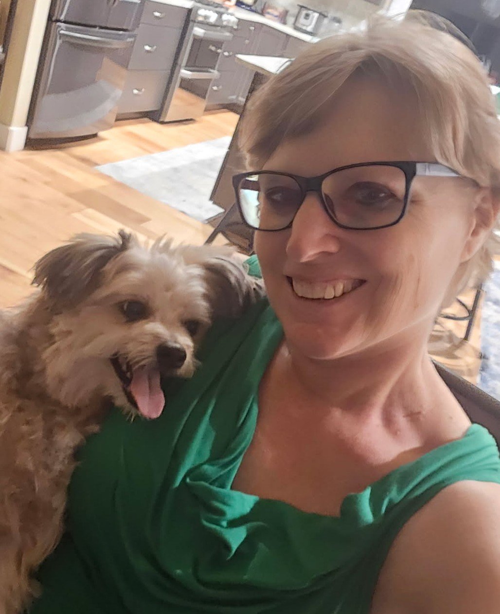 A woman with short blonde hair and glasses smiling while holding a small, fluffy dog with a tan and gray coat and an open-mouthed expression. They are in a kitchen with wooden flooring and stainless steel appliances.
