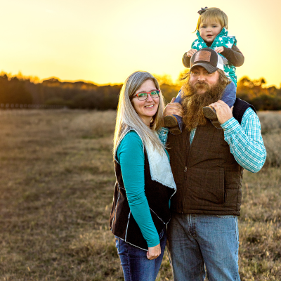 Family standing in a pasture during golden hour. Dad has daughter on his shoulders.