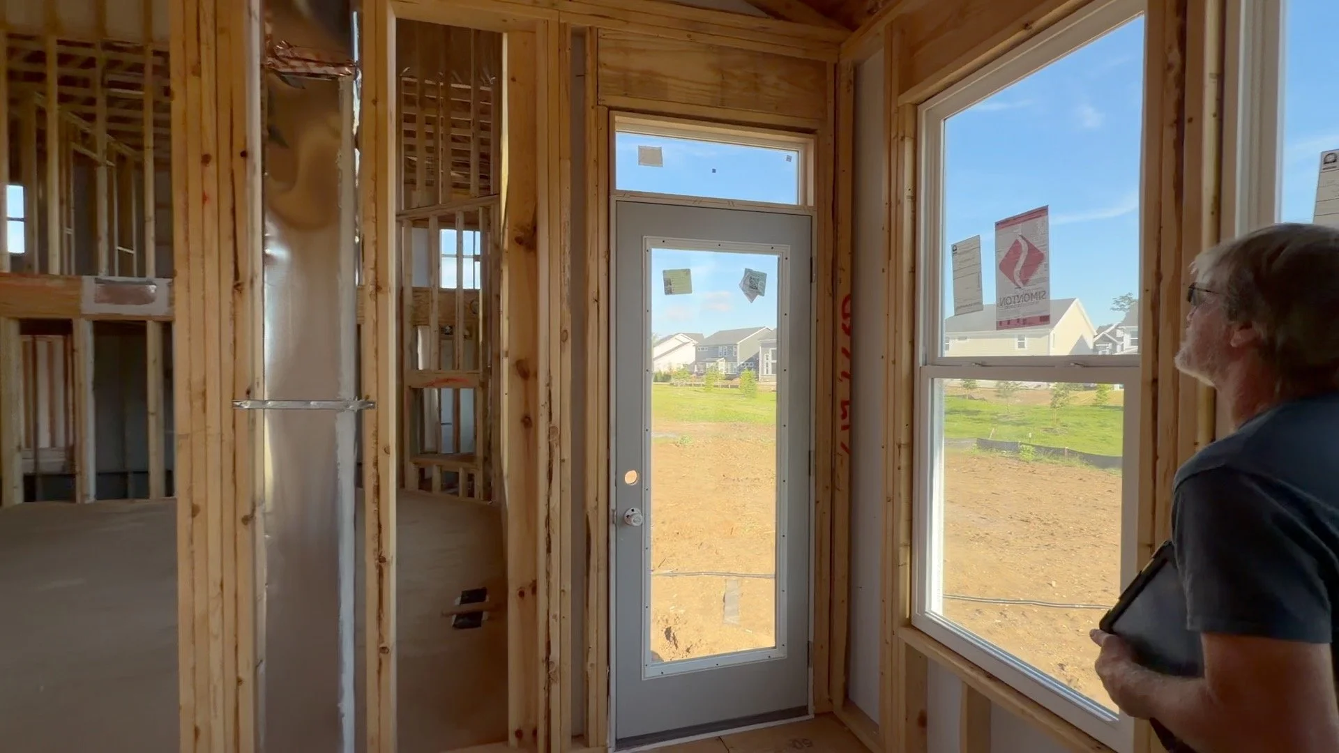 Interior view of a house under construction with exposed wooden framing, a door with a window, and a man inspecting the space, outside showing houses and a clear sky.
