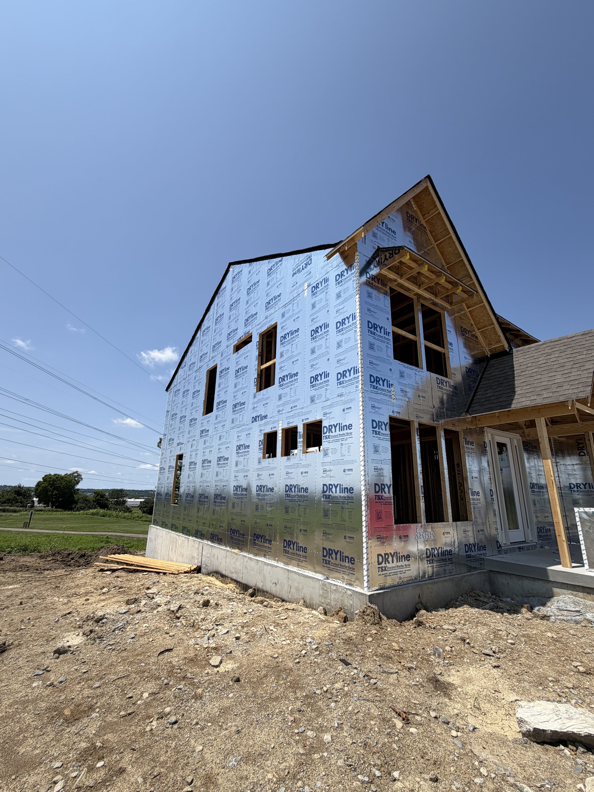A house under construction with exposed framing and a weather-resistant barrier. The house is situated on a dirt lot with construction debris and wooden planks in the foreground. The sky is clear with a few clouds and power lines are visible in the background.