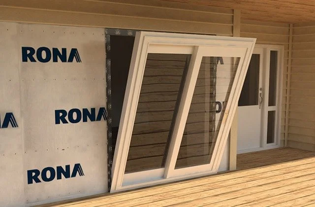 A partially installed large white-framed window leaning against a wall outside a house under construction, with a door and siding visible in the background.