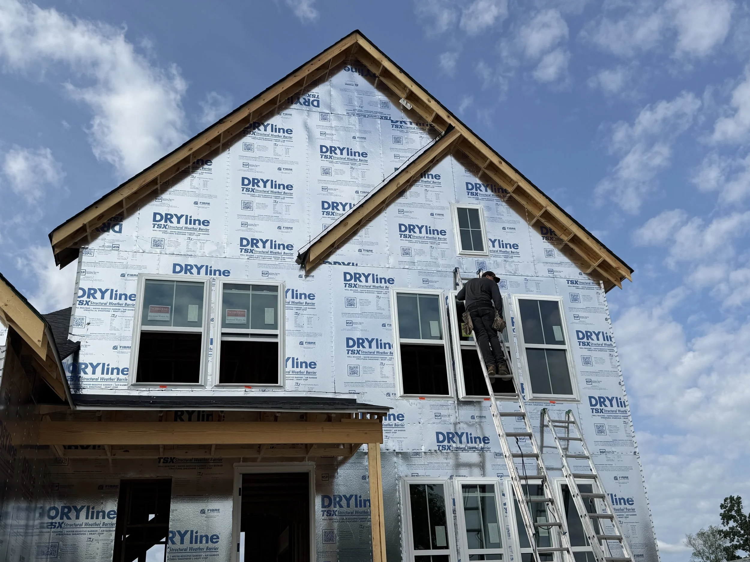 A house under construction with its exterior walls covered in a weatherproofing material labeled 'DRYline'. A person is working on the house, standing on a ladder next to the windows, with a clear blue sky and some clouds overhead.