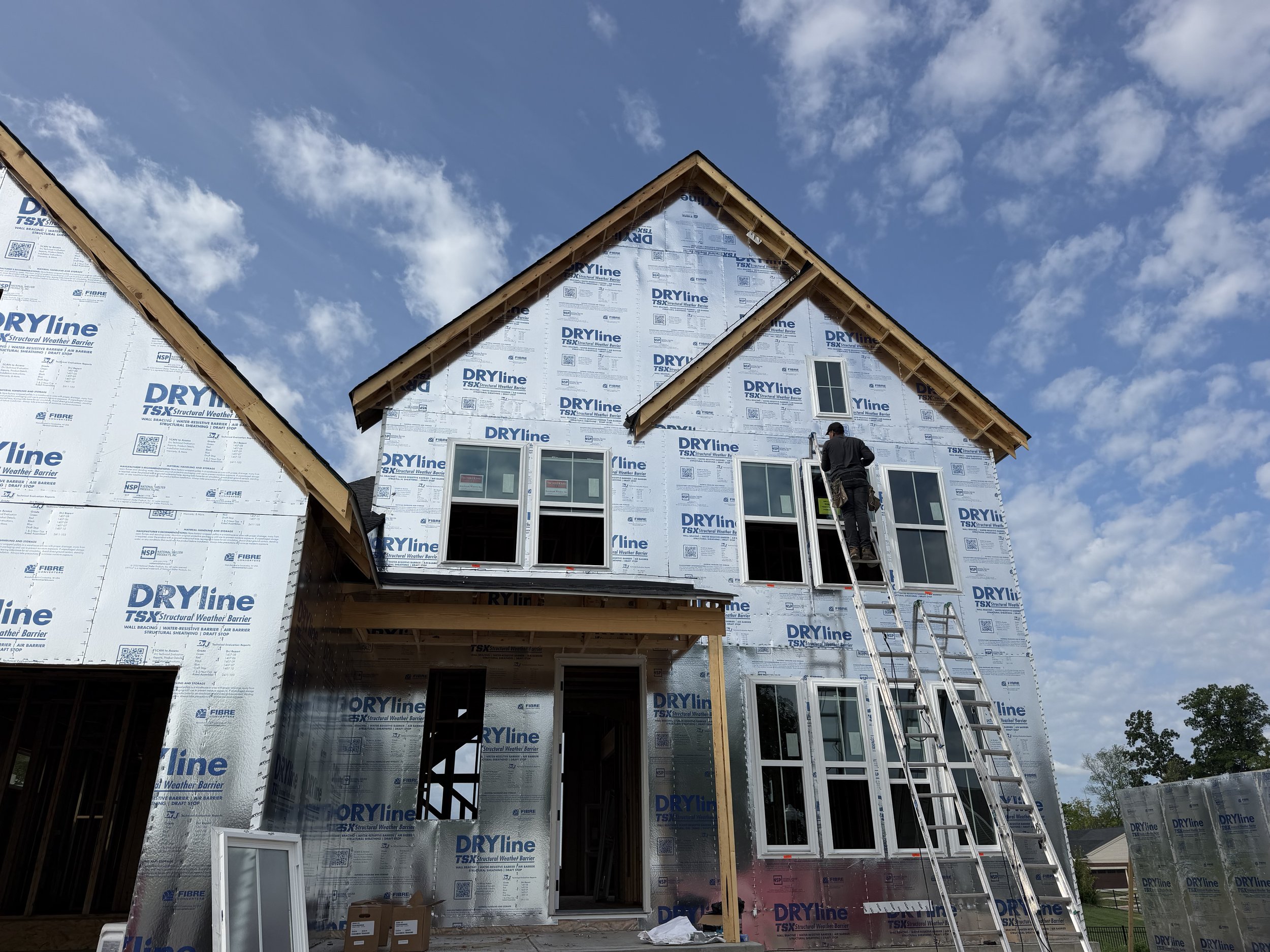 A house under construction with exterior walls covered in building wrap, with a worker on a ladder near the windows, and a partly finished roof against a background of blue sky with clouds.