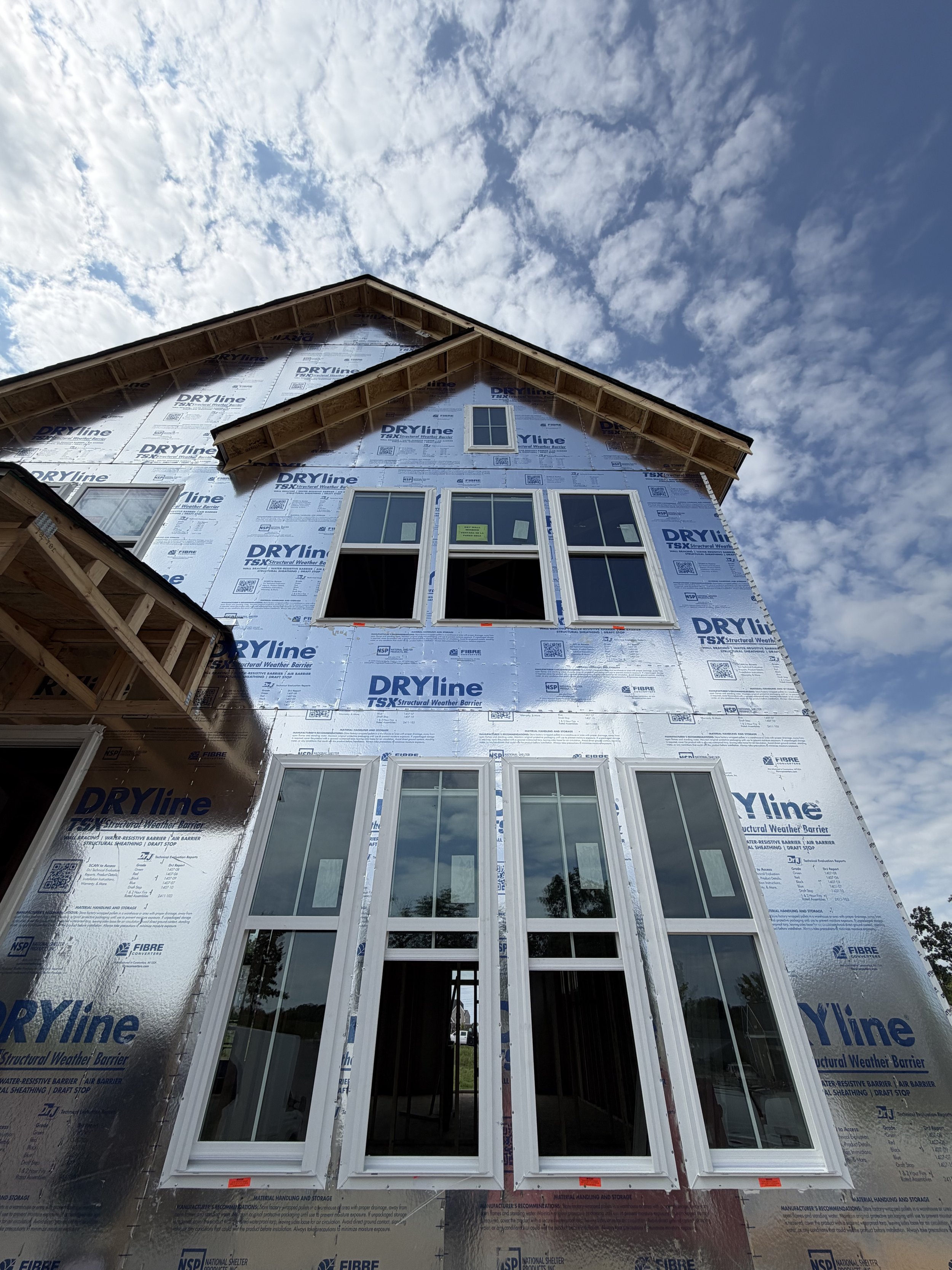 The image shows a house under construction with exterior sheathing material labeled 'DRYline' and multiple windows installed, with the sky and clouds above.
