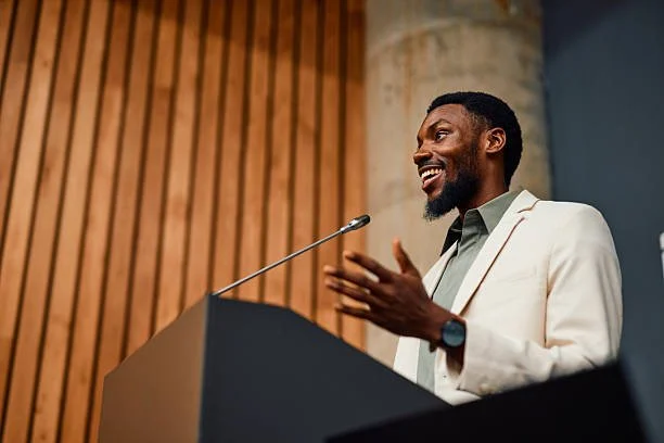 Man speaking at a podium in a suit, smiling, and gesturing with his right hand.