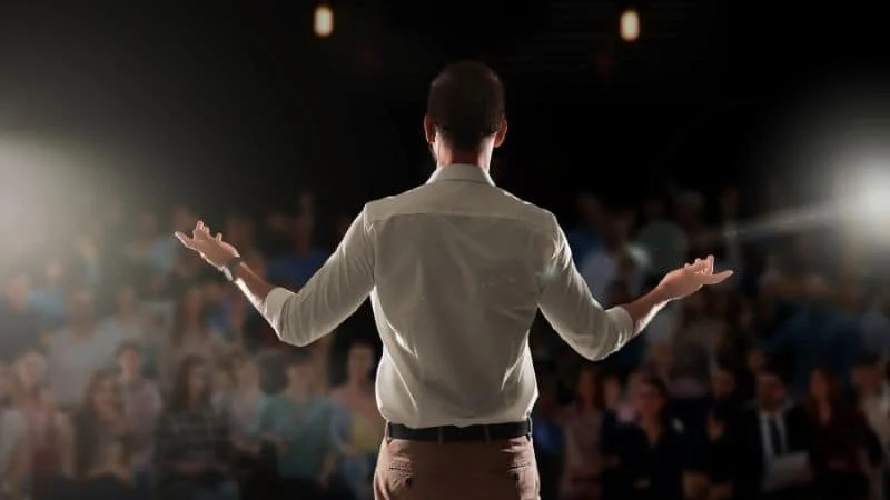 A man standing on stage with arms outstretched, facing an audience in a dimly lit room.