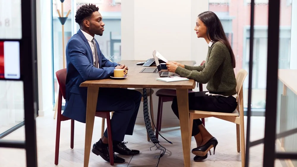 A man in a suit and a woman in a sweater and heels having a discussion at a coffee table in an office.