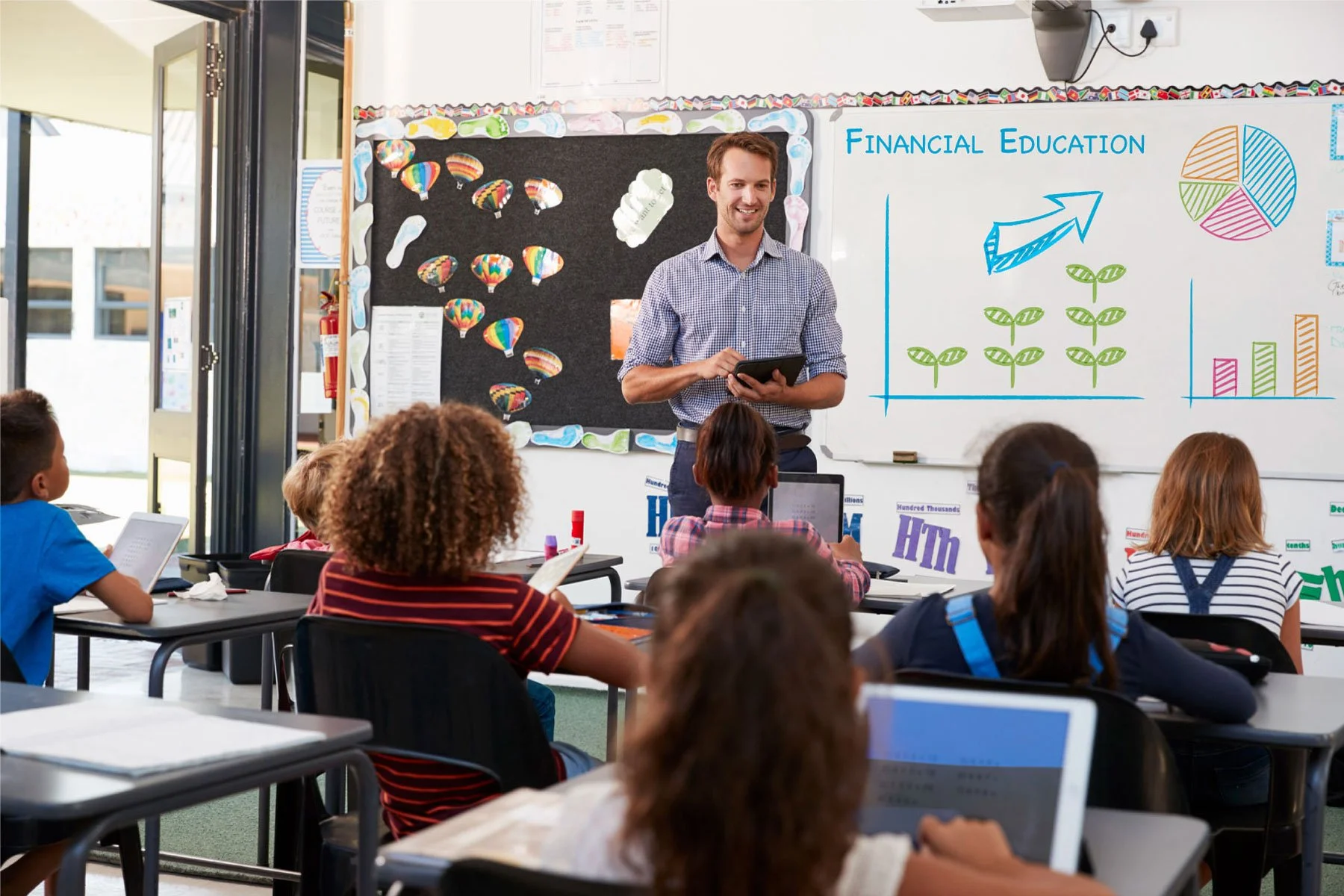 A classroom with students sitting at desks and a teacher standing at the front, teaching a lesson about financial education with diagrams on a whiteboard.