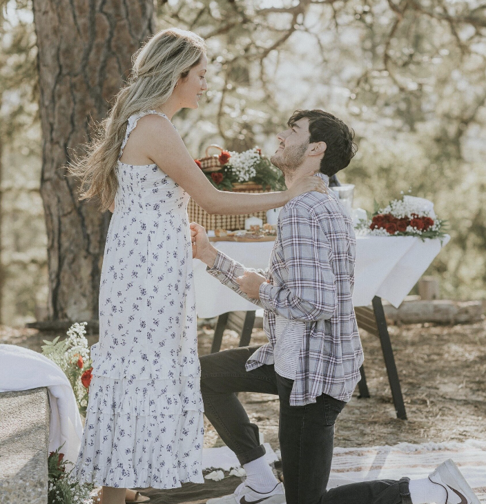 A man proposing marriage to a woman in an outdoor setting, with a table of food and decorations in the background.