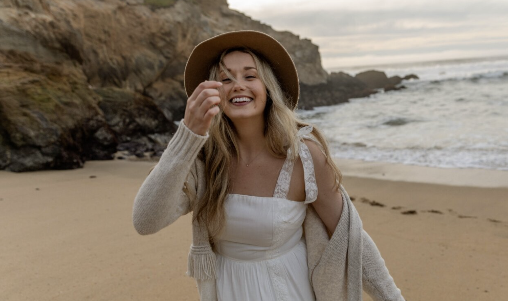 A woman with long blonde hair wearing a wide-brimmed hat, white dress, and beige cardigan smiling on a sandy beach near rocky cliffs with ocean waves in the background.