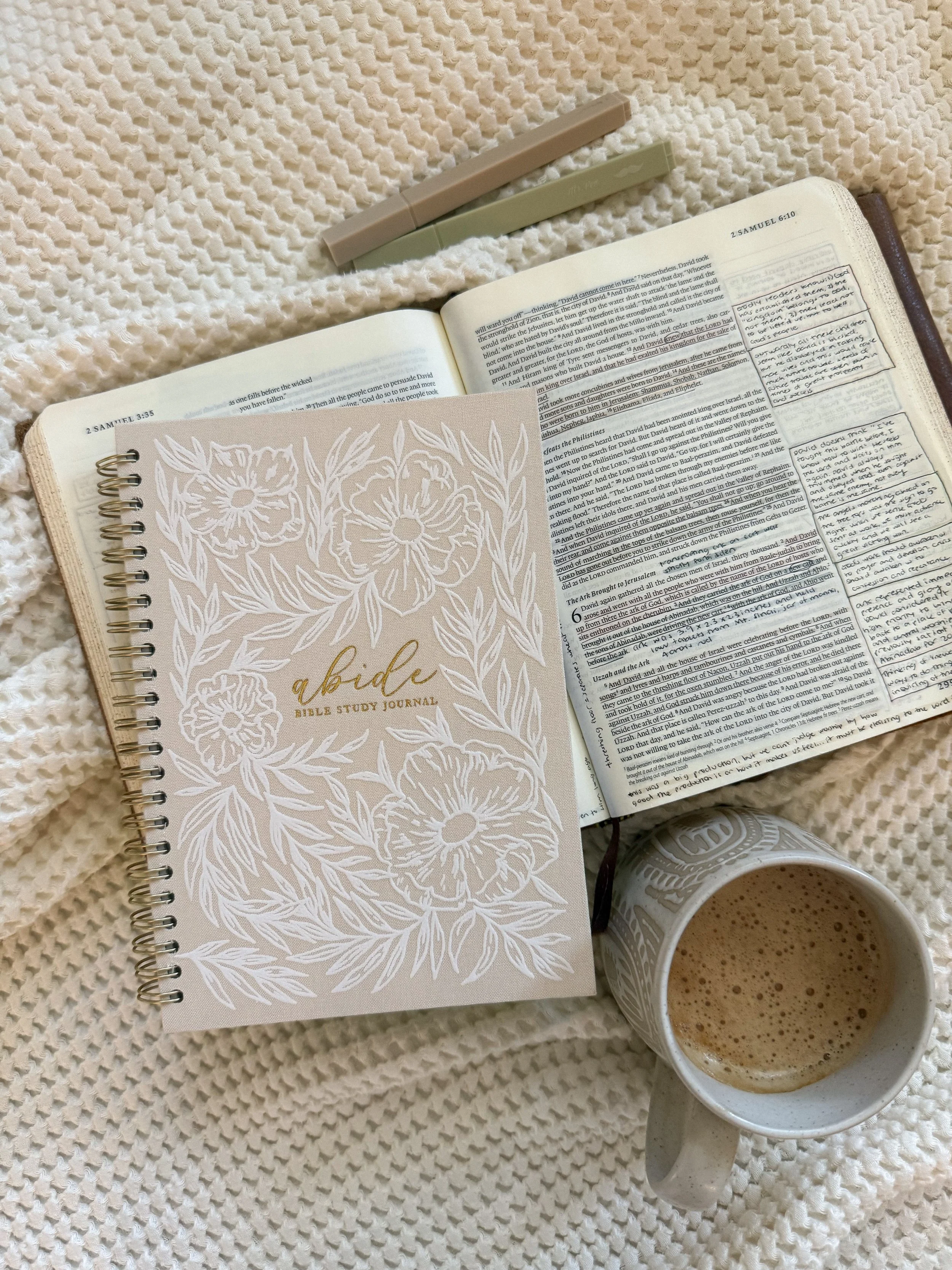 An open Bible with highlighted verses, a spiral notebook with a floral design titled 'a bible,' a cup of coffee, and two pens on a textured white blanket.