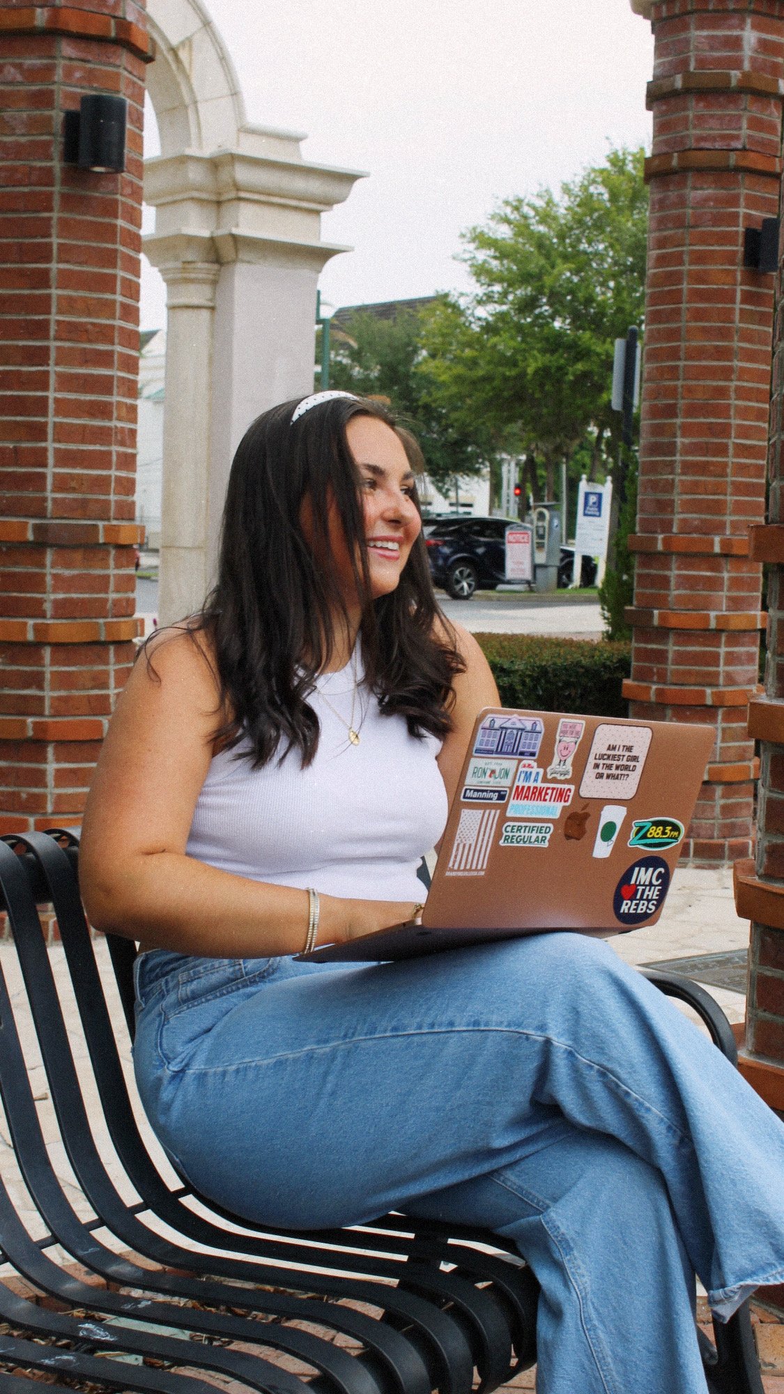 A young woman with long dark hair sitting outside on a bench with a laptop covered in stickers, smiling, wearing a white tank top and blue jeans, in front of a brick and stone archway with trees and a parking lot in the background.