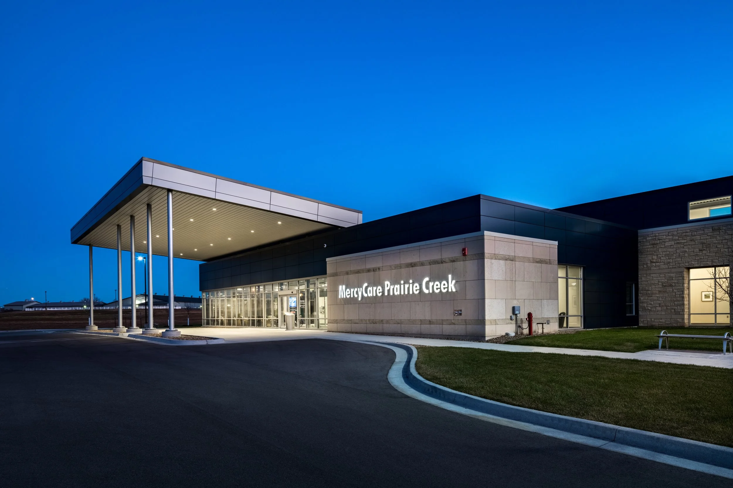 Modern medical facility building named MercyCare Prairie Creek at dusk, with a clean and sleek design, illuminated signs, and a well-maintained parking area.