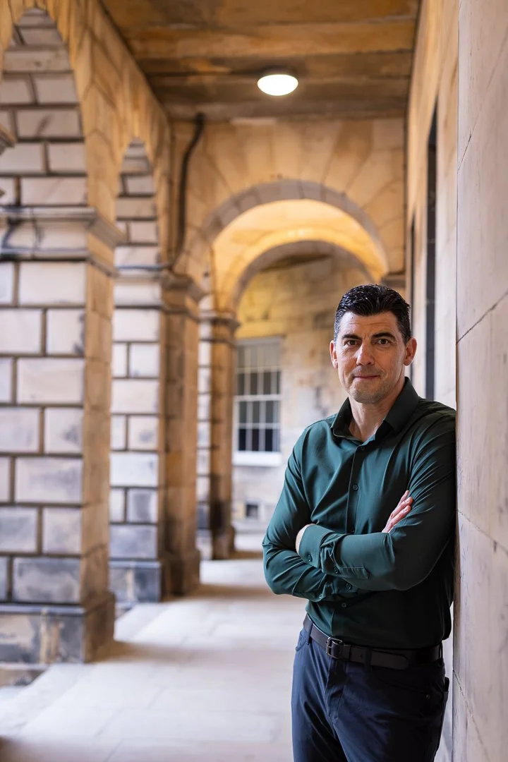 Craig in a dark green shirt, standing with arms crossed in an arched, historic stone building corridor.