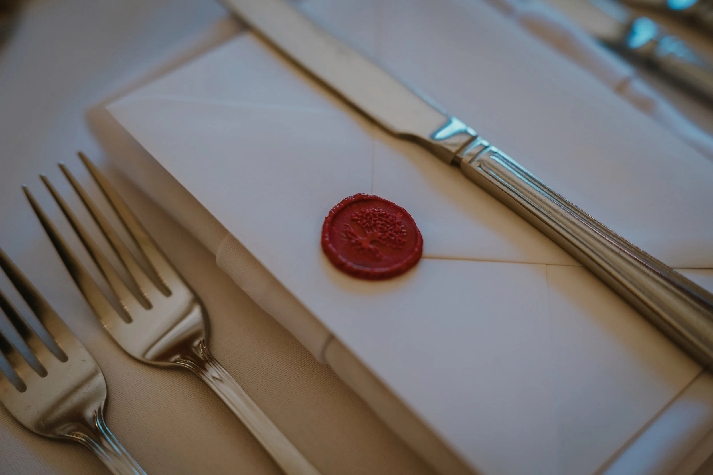 Silver knife with an engraved handle resting on a closed envelope sealed with red wax featuring a carved emblem, and a silver fork beside it, all placed on a white surface.