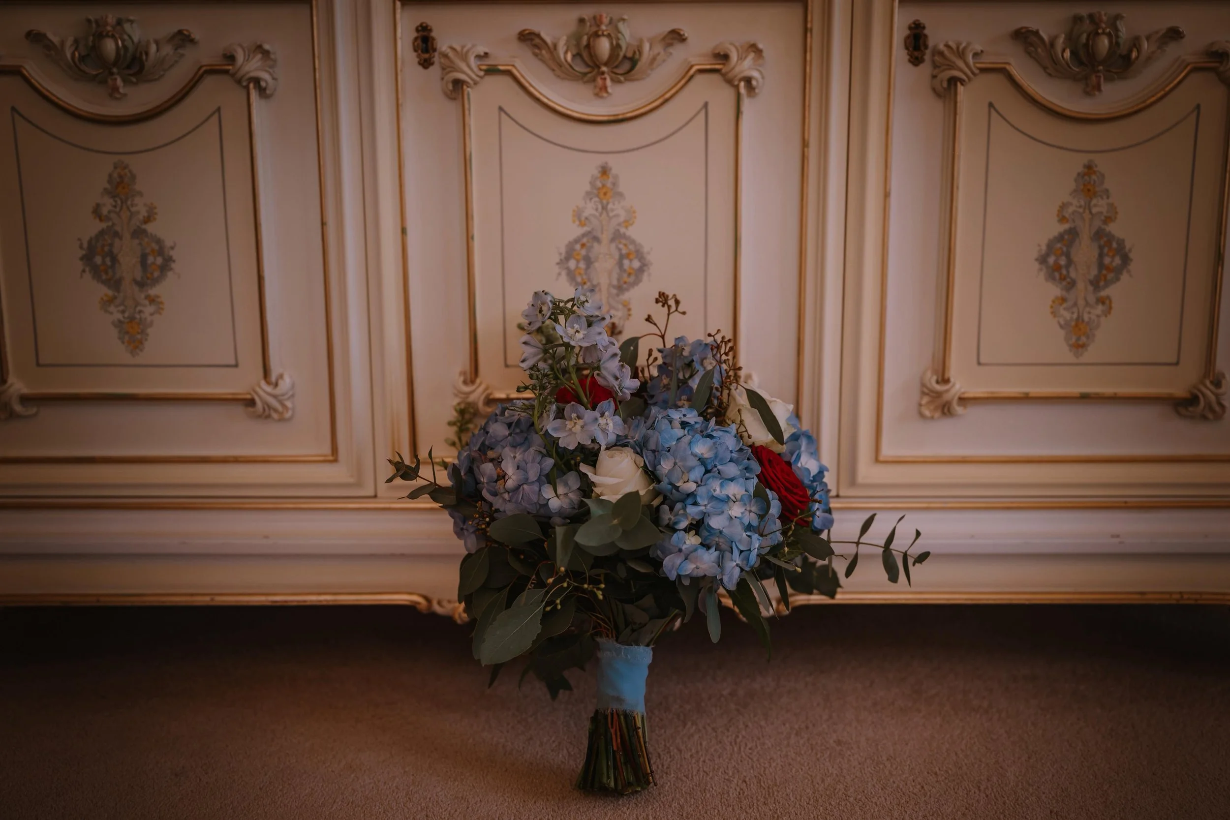 A large bouquet of blue, white, and red flowers, including hydrangeas and roses, arranged in a glass vase in front of a vintage, ornate cream-colored cabinet with gold accents.
