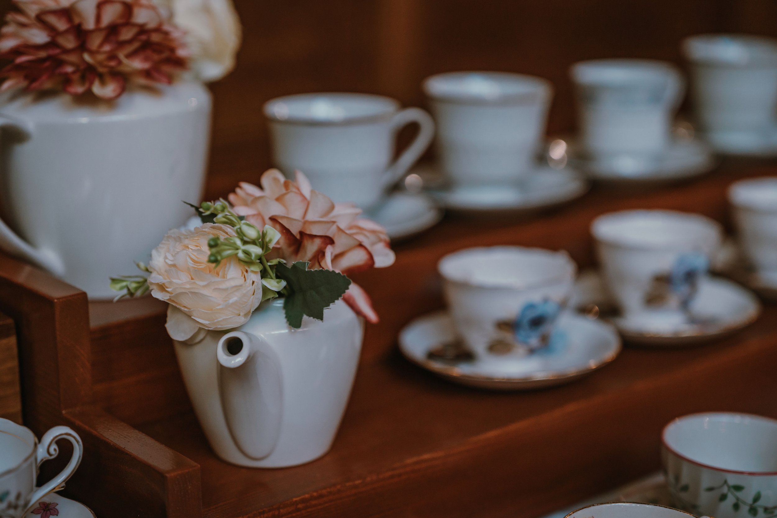 A white ceramic teapot with pink and cream artificial flowers inside it, placed on a wooden shelf. In the background, there are several white teacups stacked on saucers, some with floral patterns, on the same shelf.